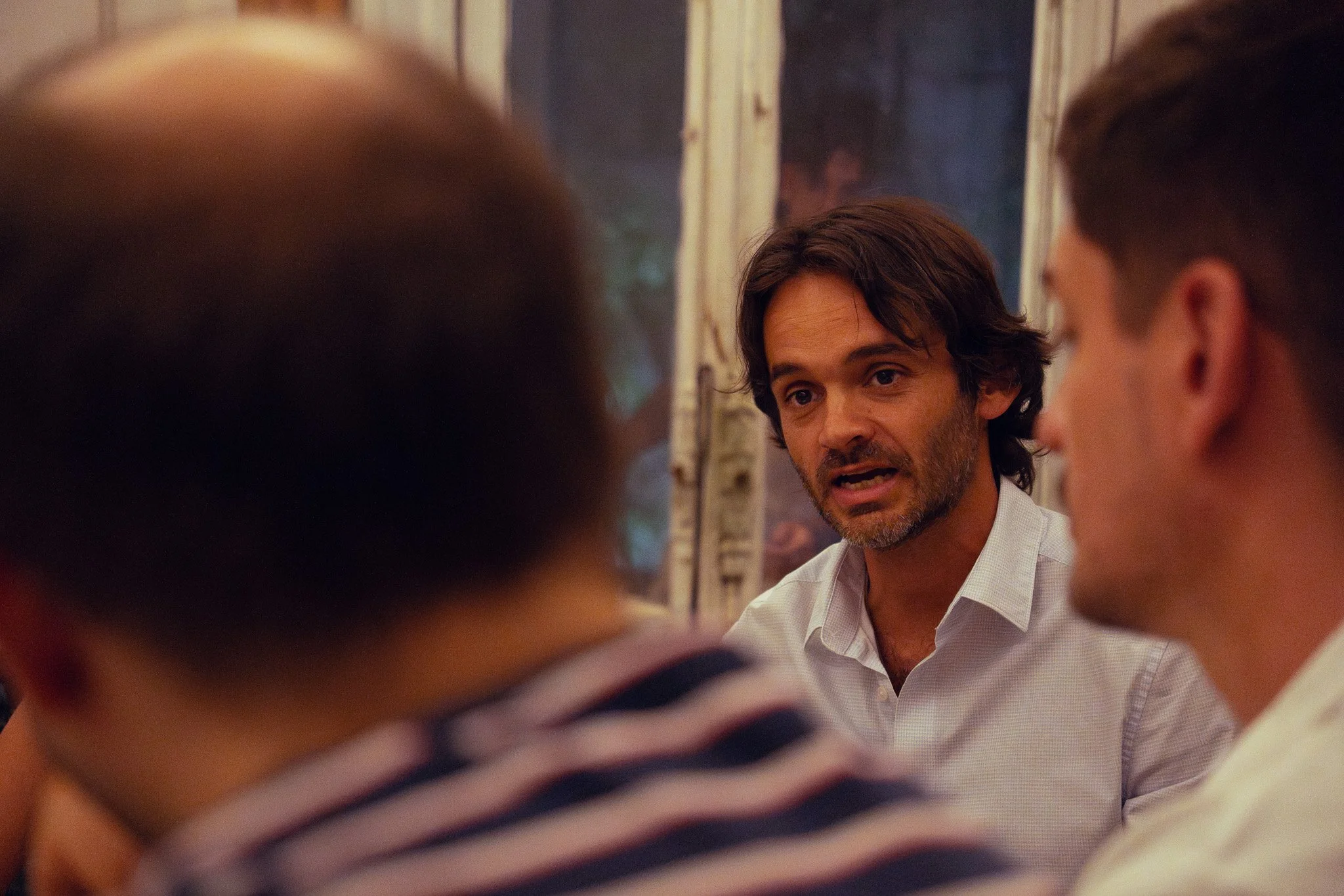 Un hombre con barba y cabello castaño oscuro, vestido con una camisa blanca, hablando con un grupo de personas en un entorno interior.