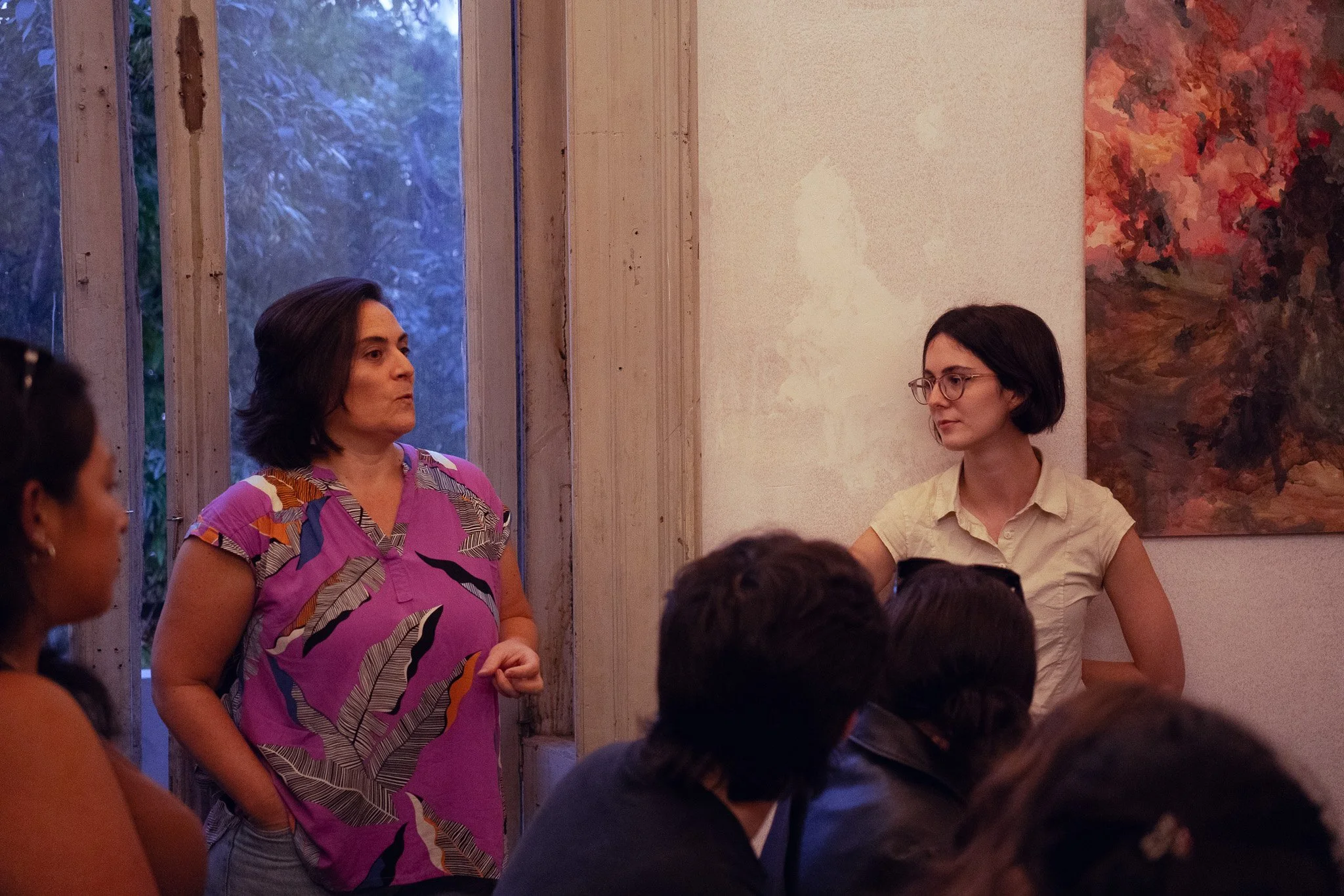 Una mujer con gafas y camisa beige está de pie junto a una pared, escuchando a otra mujer con camiseta morada con estampado de hojas que habla frente a un grupo de personas. Están en un espacio interior con una ventana de madera y un cuadro de colore