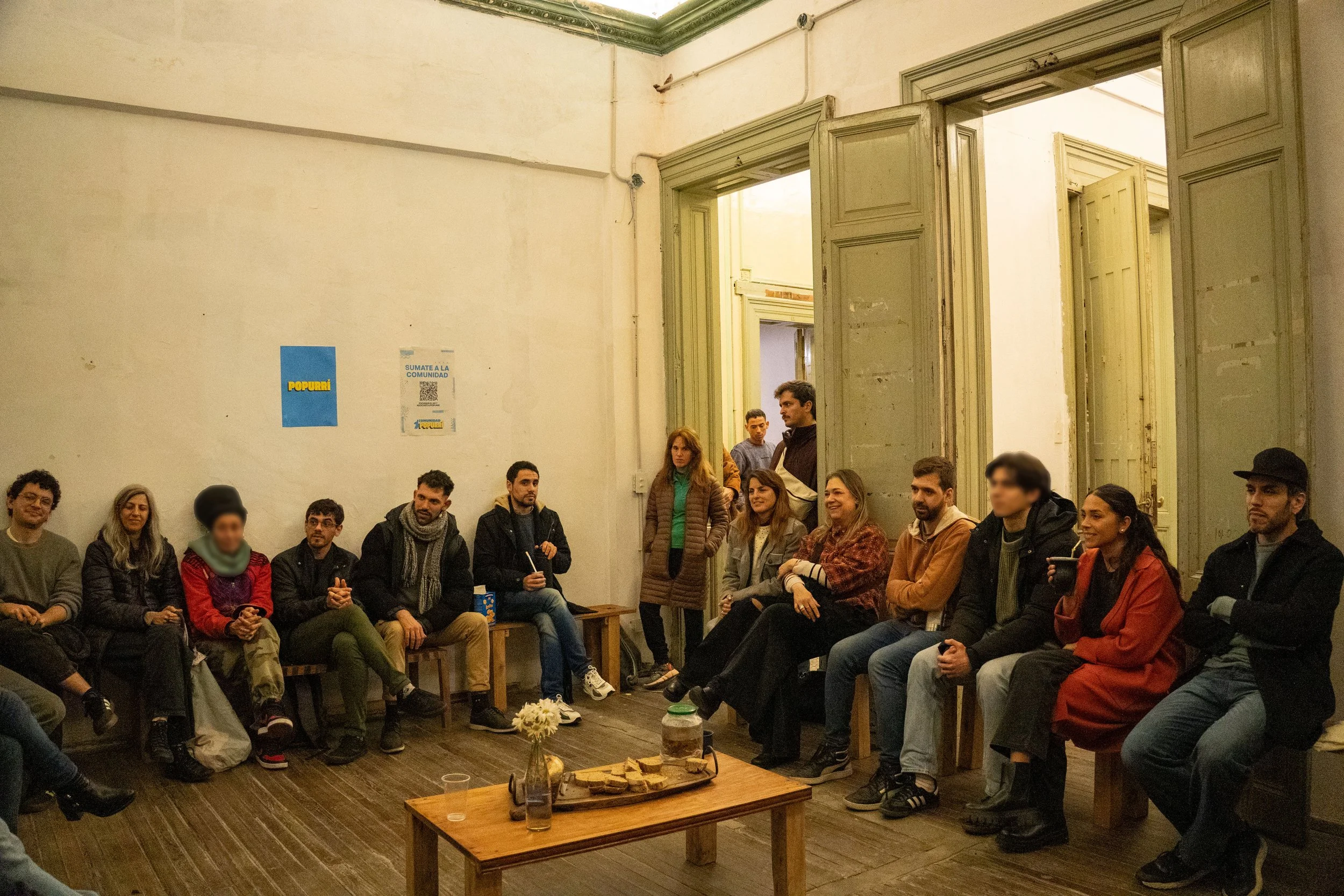 Un grupo de personas sentado en una sala, participando en una reunión o evento. En el centro hay una mesa con comida y bebidas, y en las paredes hay anuncios y posters.