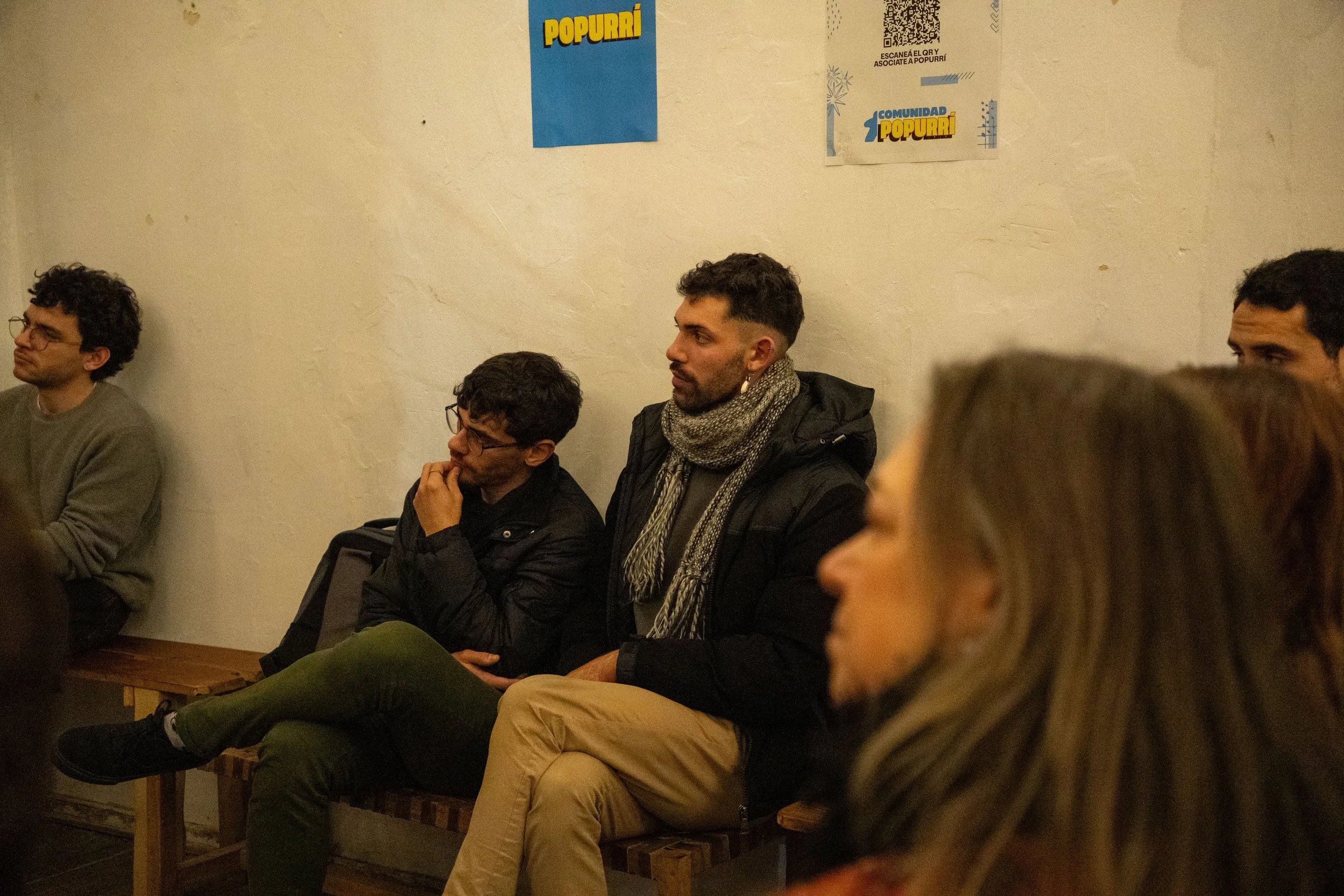 Varias personas sentadas en una sala, algunas mirando hacia adelante, en un ambiente interior con paredes claras y carteles en la pared que dicen 'Popurri'.