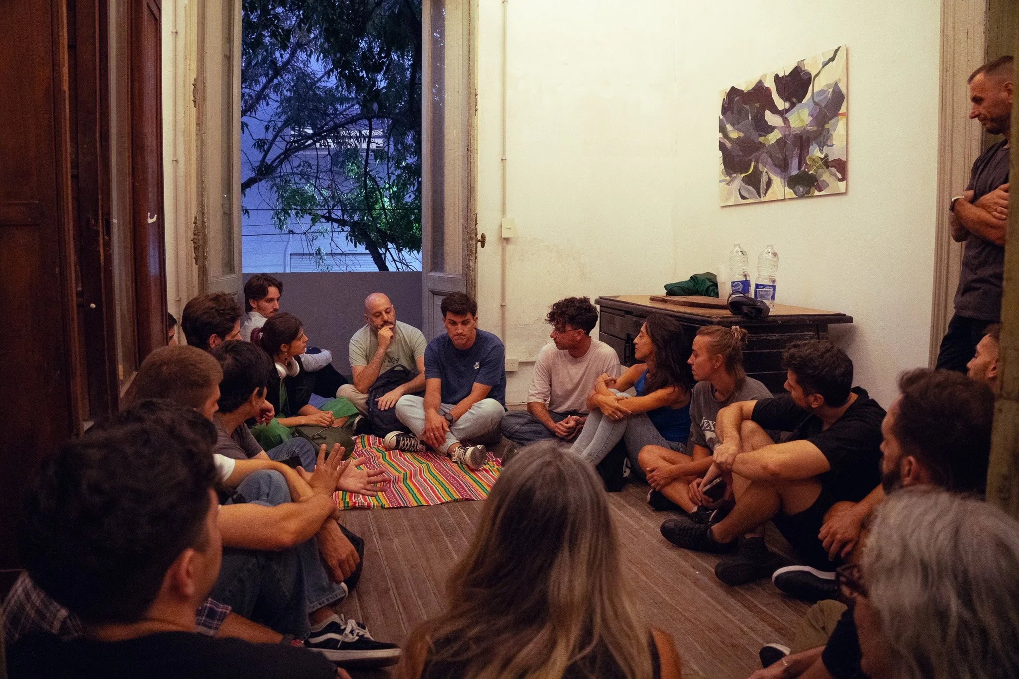 Grupo de personas sentadas en círculo en una sala, participando en una discusión o reunión, con una ventana y arte en la pared.