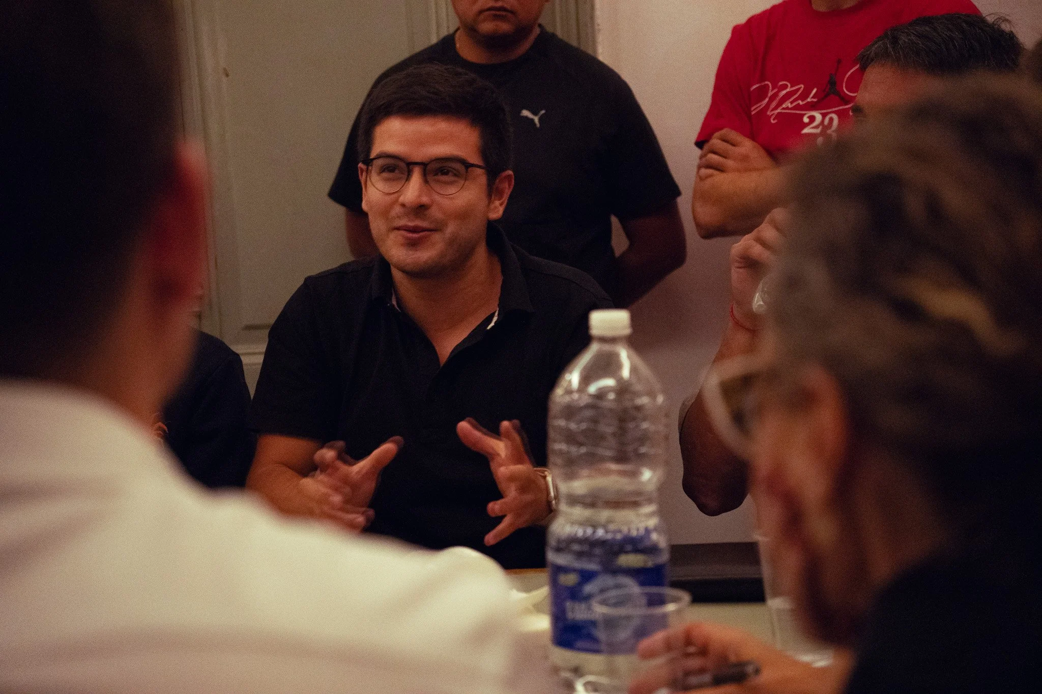 Hombre con gafas y camiseta negra hablando en una reunión, rodeado de varias personas.