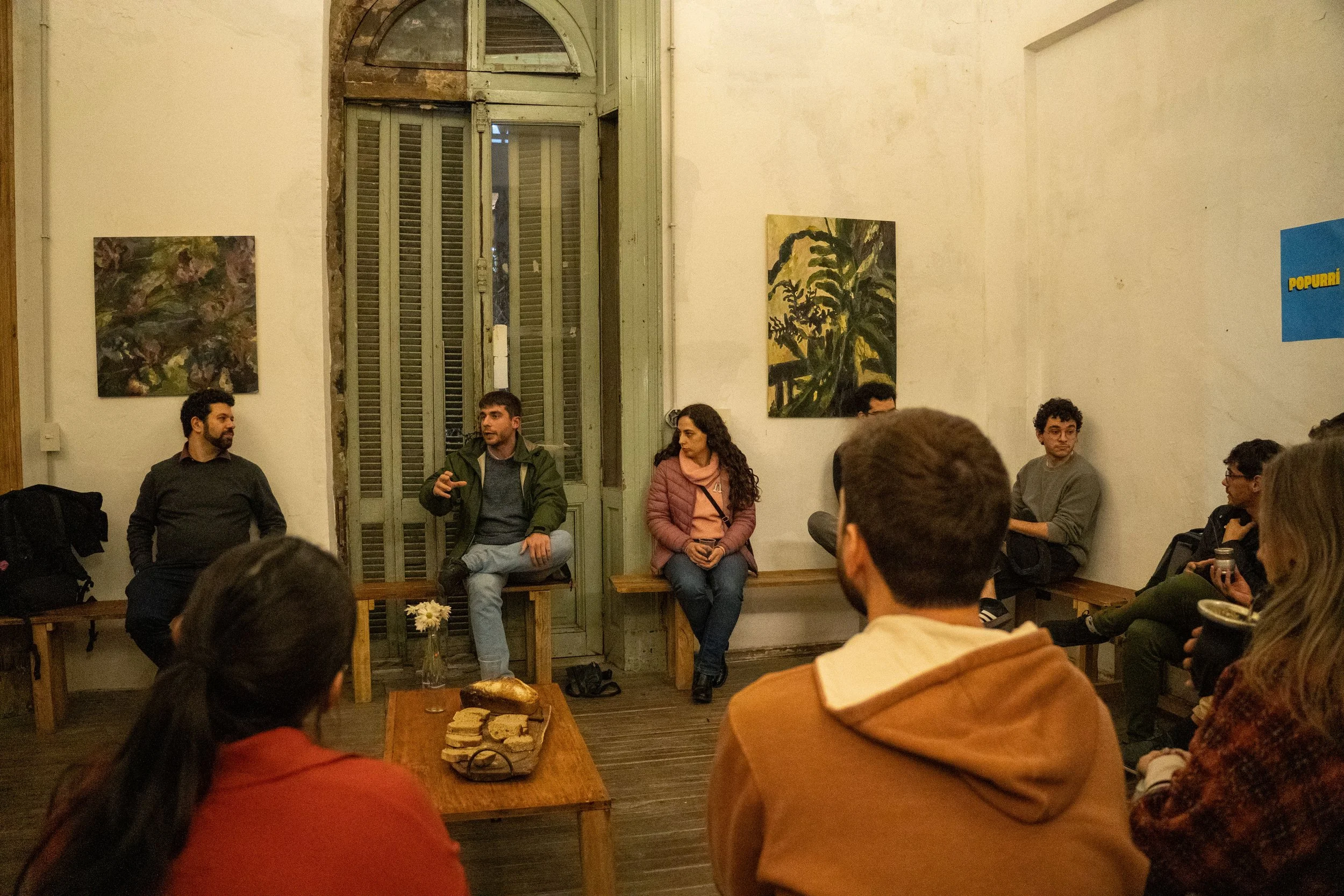 Personas sentadas en un círculo en una sala de arte o reuniones, participando en un diálogo, con pinturas colgadas en la pared y una mesa con comida y bebidas en primer plano.