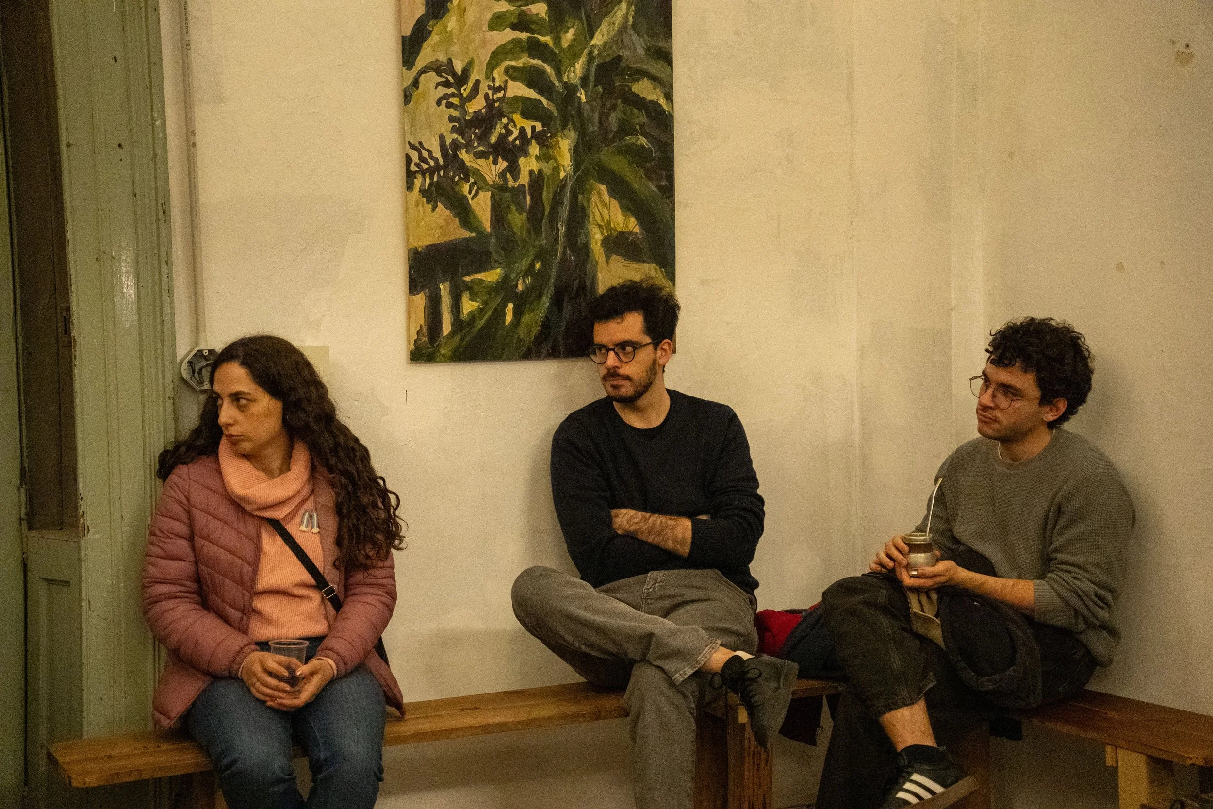 Tres personas sentadas en un banco de madera y dos más en una banca contra la pared en un interior, con un cuadro pintado en la pared detrás de ellos.