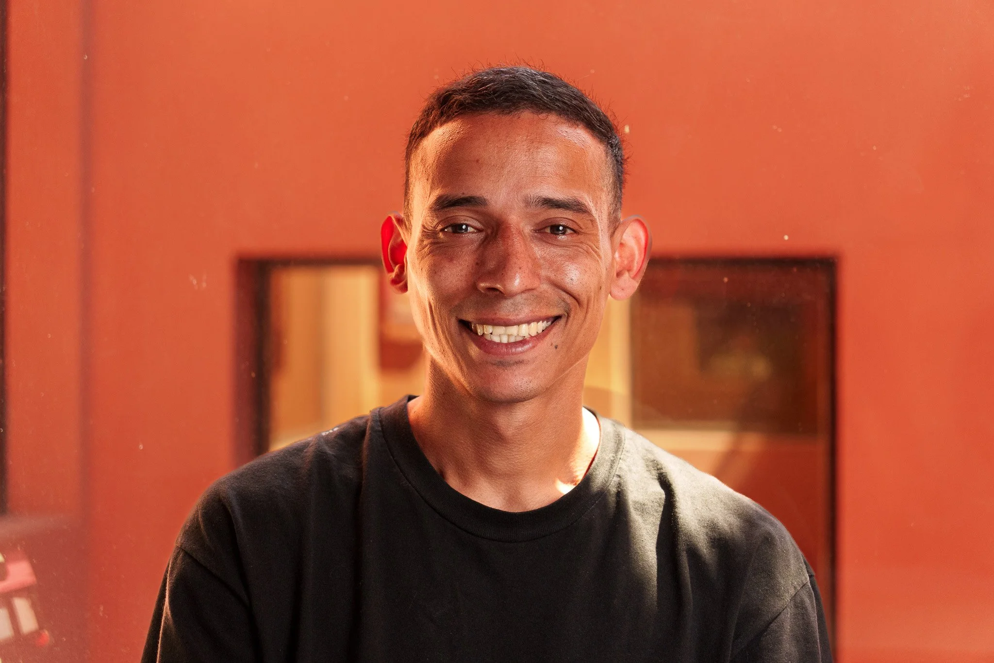 Hombre sonriendo en un ambiente con paredes naranjas y un espejo en el fondo.