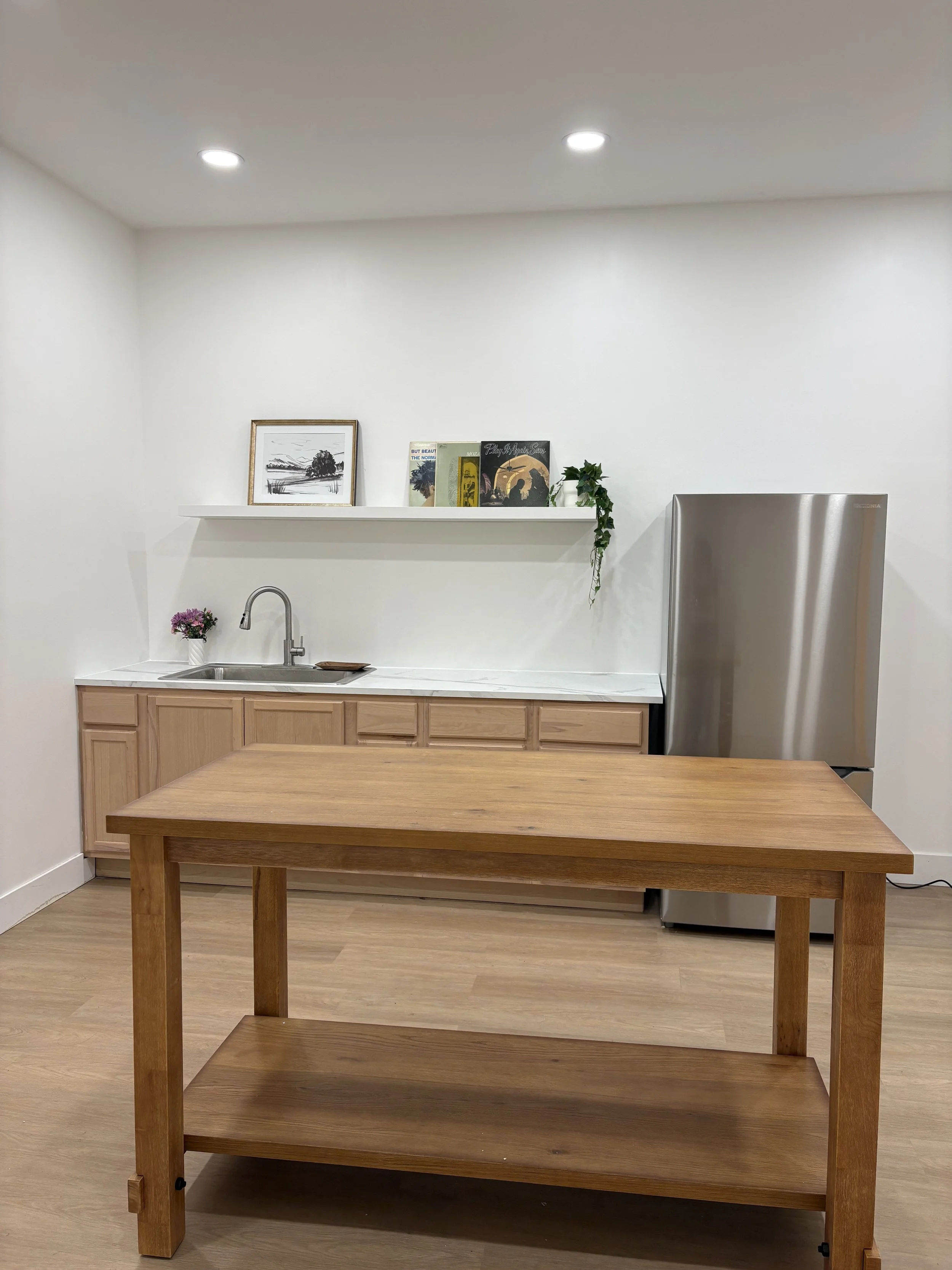Minimalist kitchen with white walls, wooden floor, stainless steel refrigerator, wooden table, and a white countertop with a sink. A shelf above holds framed artwork, books, and a small potted plant.