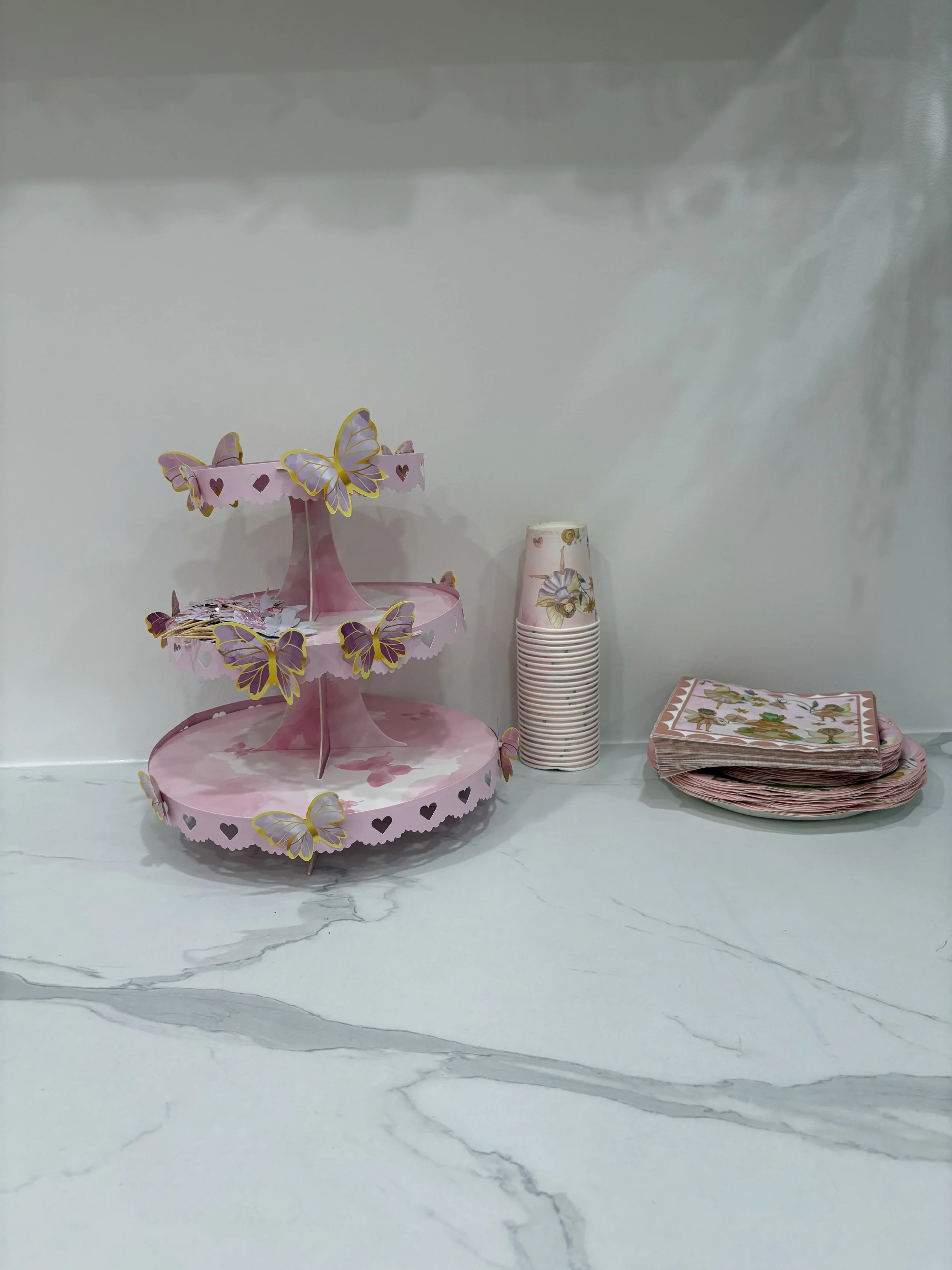 Pink tiered cupcake stand with butterfly decorations, a stack of paper cups with floral designs, and a stack of paper napkins with a floral pattern, on a white marble countertop.