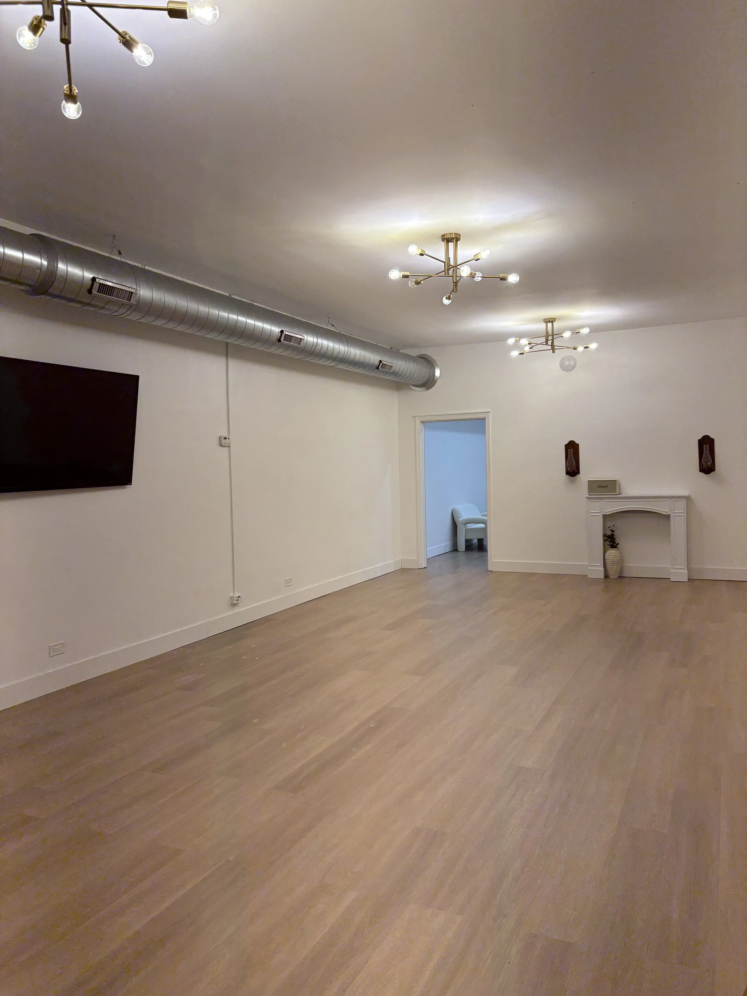 An empty room with beige flooring, white walls, and modern ceiling lights. There is a flat-screen TV mounted on the wall, an exposed industrial duct running along the ceiling, and a doorway leading to a smaller room with a white chair. There are decorative wall sconces and a small shelf with a vase and plant.