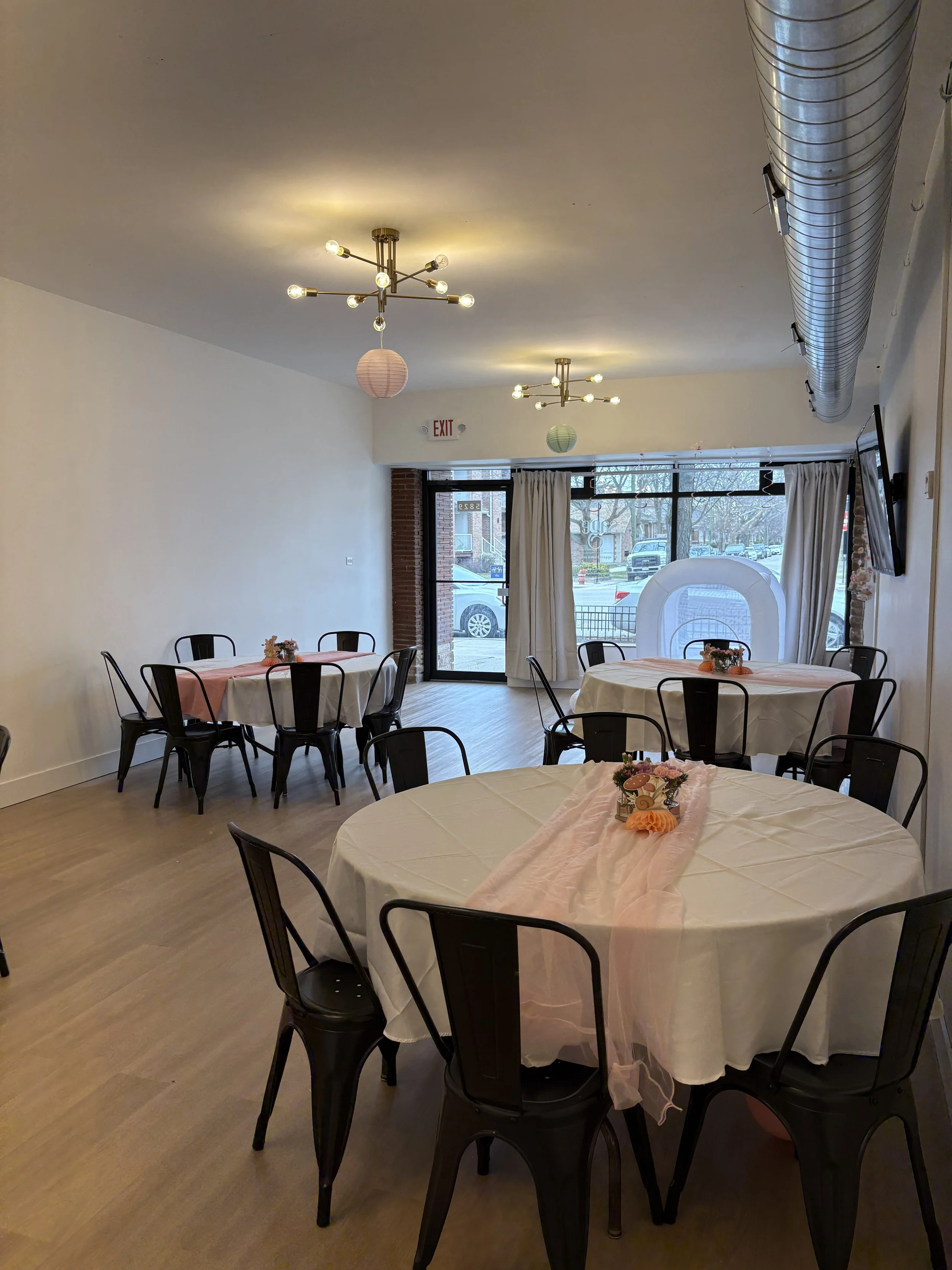Event space with round tables decorated with pink and white tablecloths and floral centerpieces, black metal chairs, ceiling mounted modern chandeliers, and floor-to-ceiling windows with white curtains.