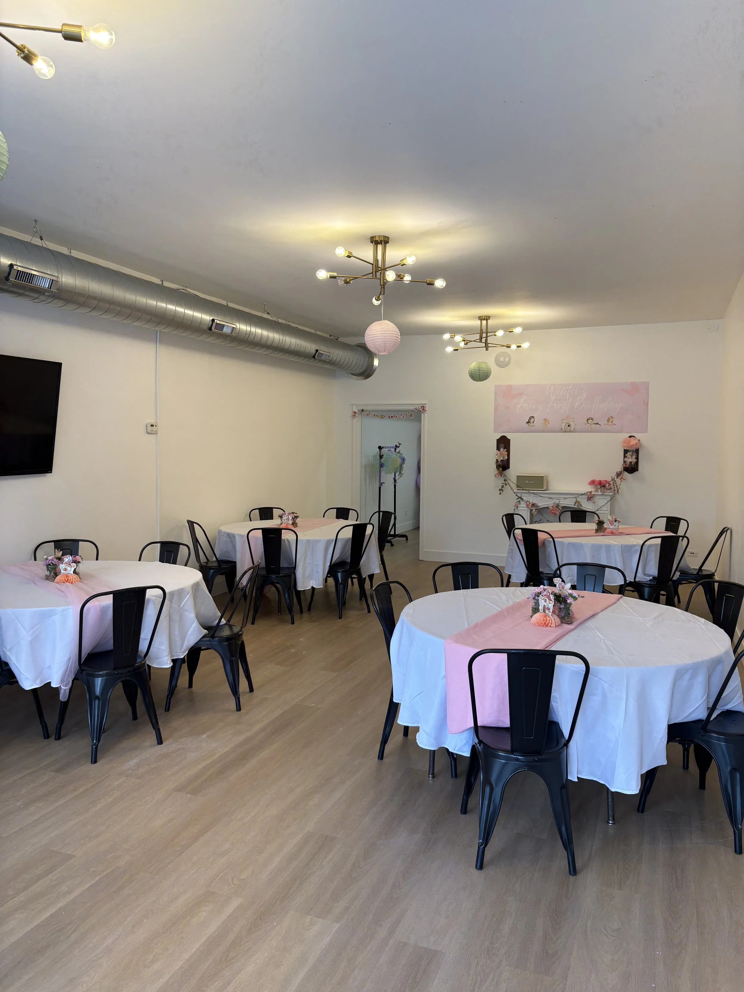 Event space decorated with round tables covered with white tablecloths and pink runners, each with floral centerpieces, black chairs, ceiling lights, paper lanterns, and a pink banner on the wall reading "Happy Wedding & Party" with floral decoration