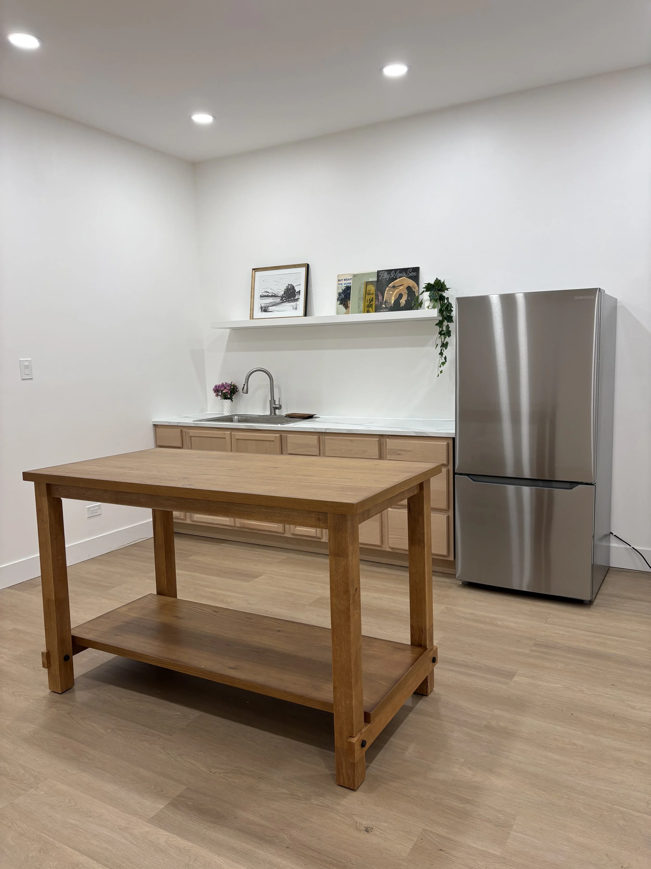 Minimalist kitchen with wooden table, white cabinets, stainless steel refrigerator, white walls, and framed artwork on a shelf.