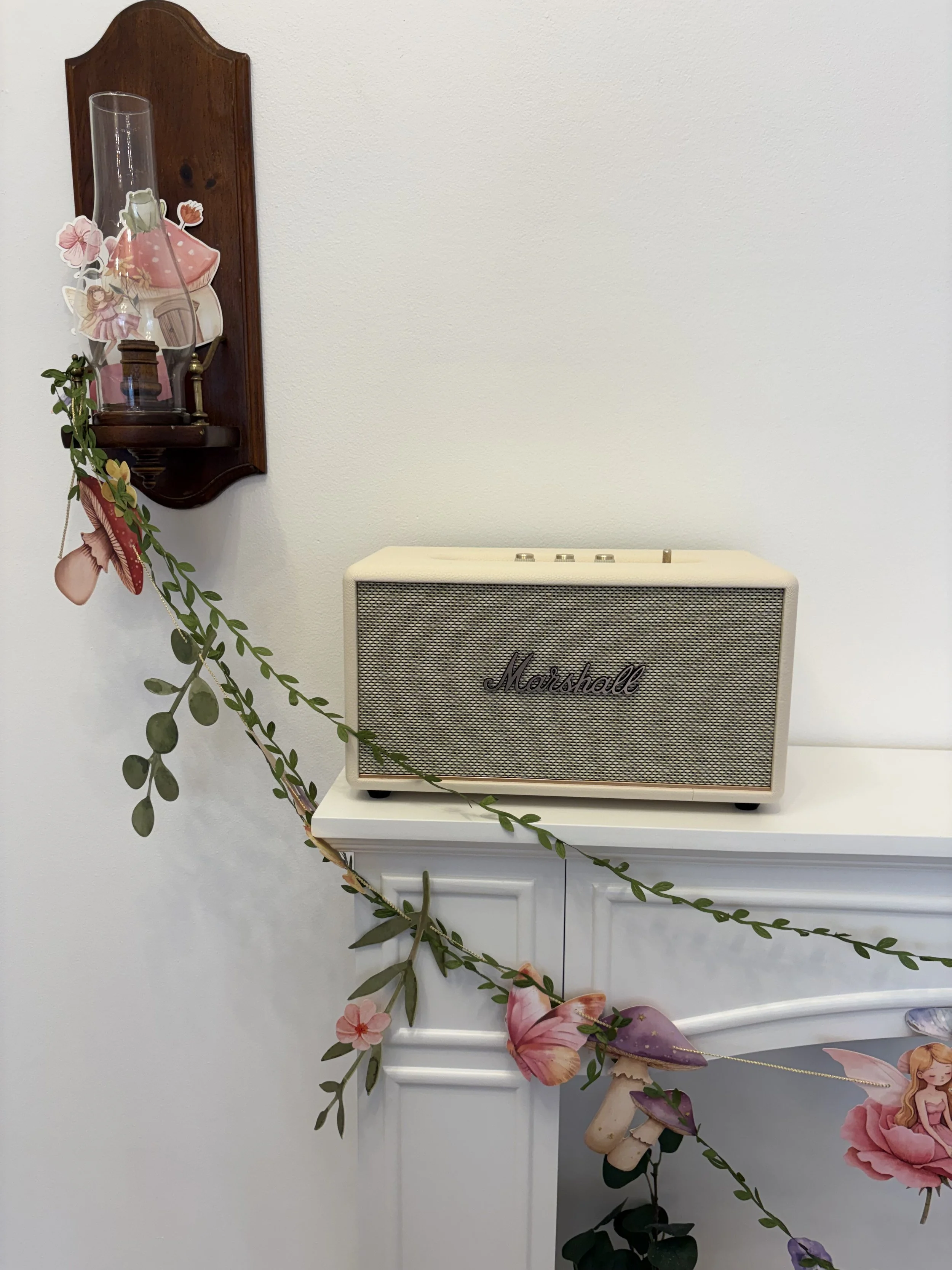 A white Marshall speaker on a white surface decorated with a garland of leaves, pink flowers, a mushroom, and fairy cutouts hanging from a wall-mounted wooden shelf, with a white wall in the background.