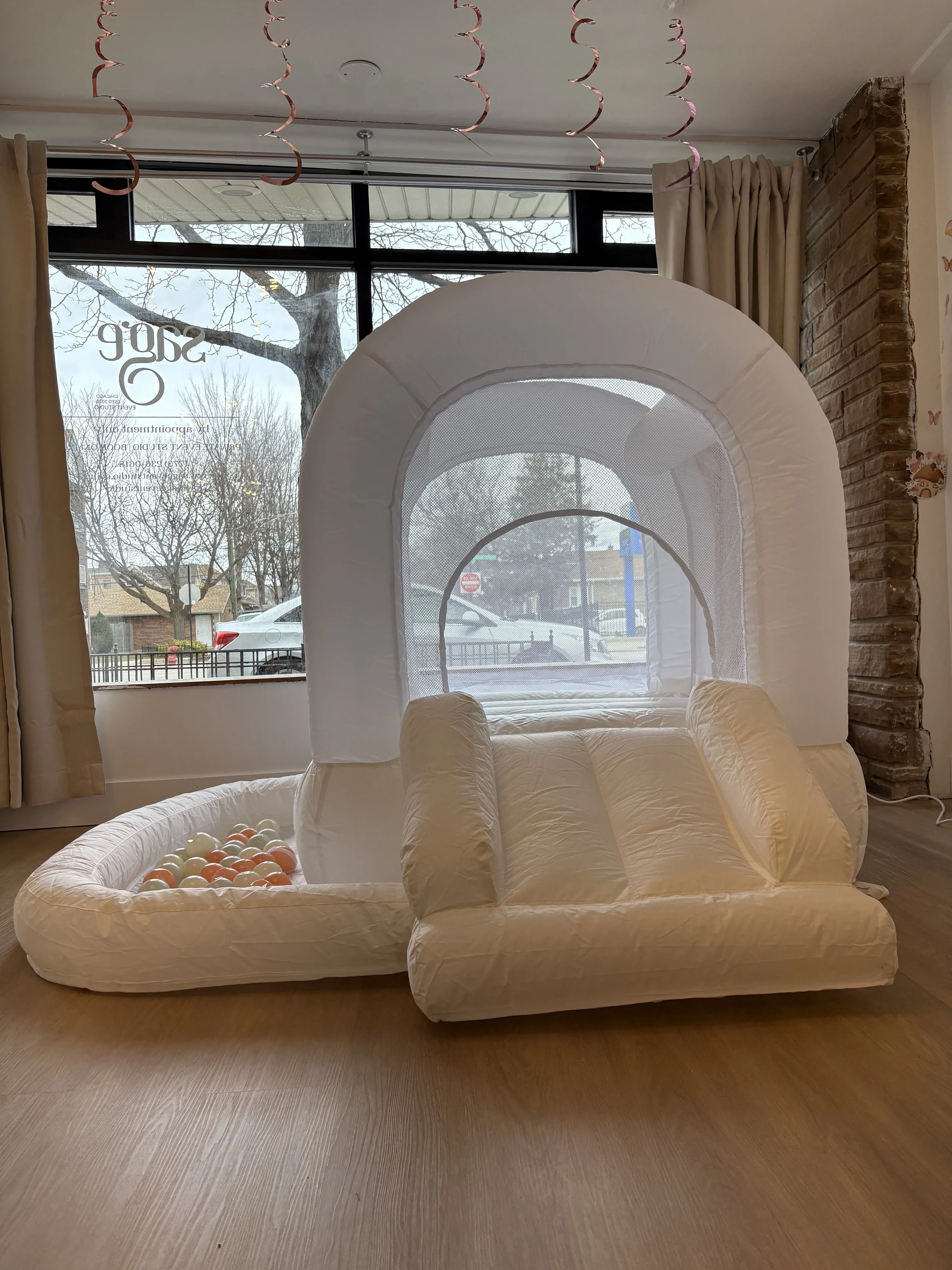 Inflatable indoor ball pit and playhouse with mesh windows, next to a large front window with curtains, in a room with wooden flooring and brick wall accents.