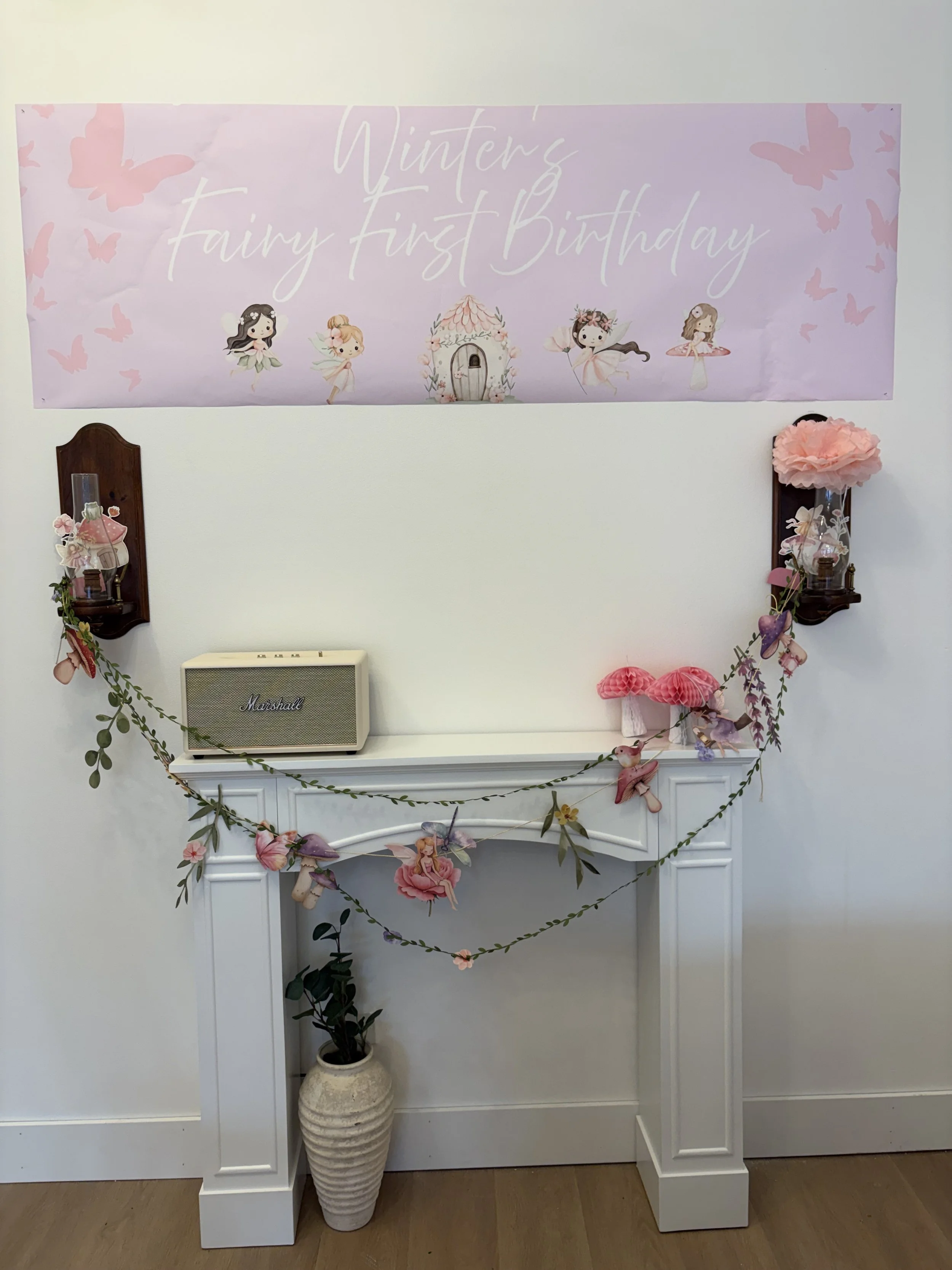 Decorative setup for a child's first birthday party with fairy theme, pink and purple accents, fairy illustrations on a banner, and a white fireplace mantle adorned with paper flowers, fairy cutouts, and a potted plant.