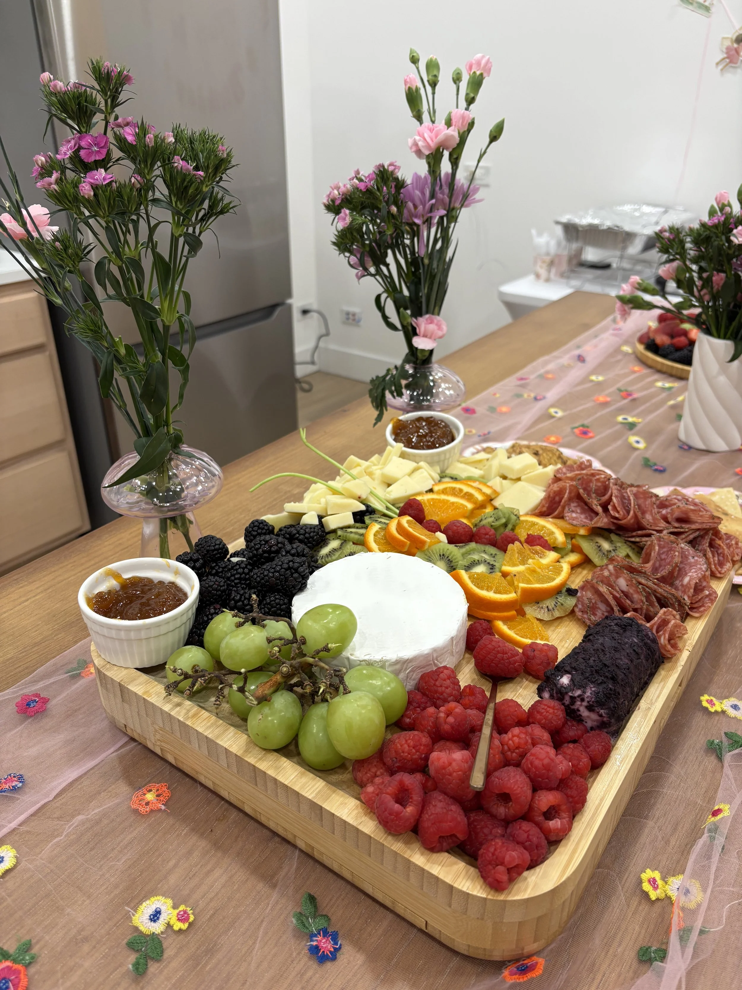 A charcuterie board with assorted fruits and meats on a wooden table with flower vases and decorative tablecloth.