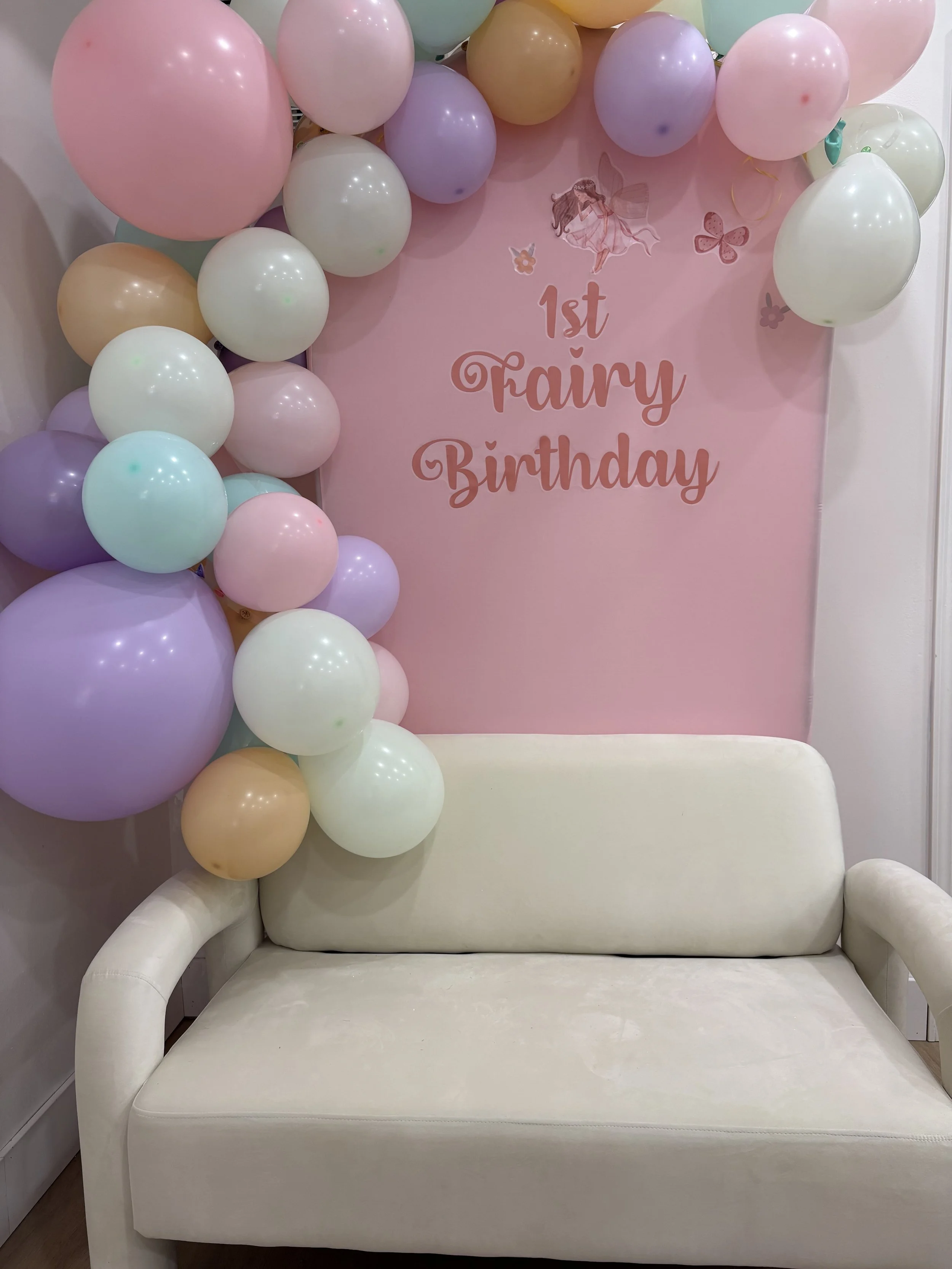 Decorative setup for a child's birthday with pastel balloons surrounding a pink backdrop that reads '1st Fairy Birthday' and features a fairy illustration, with a white cushioned sofa in front.
