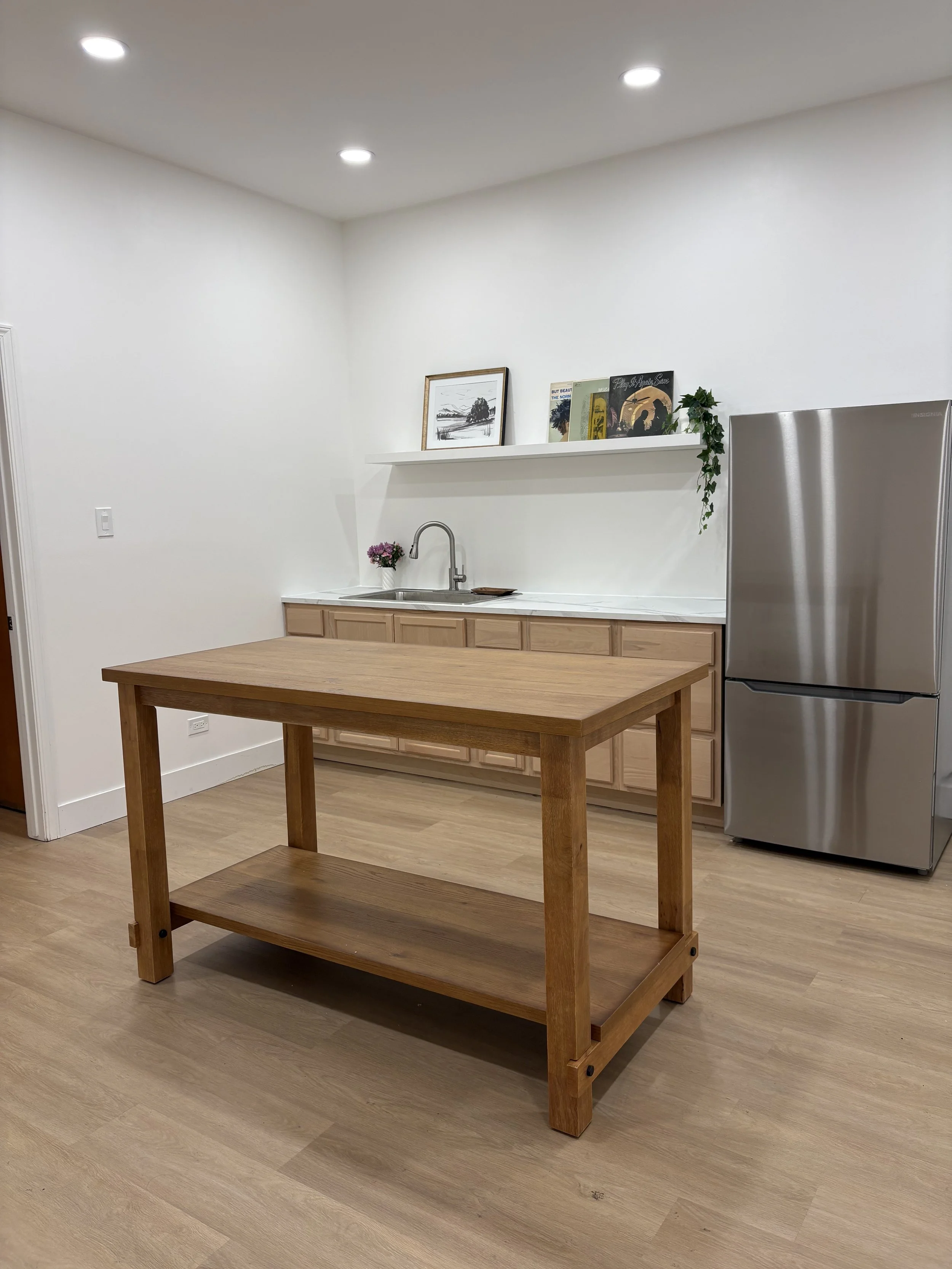 A minimalist kitchen with white walls, ceiling lights, a wooden table, a white countertop, a small sink, a stainless steel refrigerator, and a shelf with framed pictures and books.