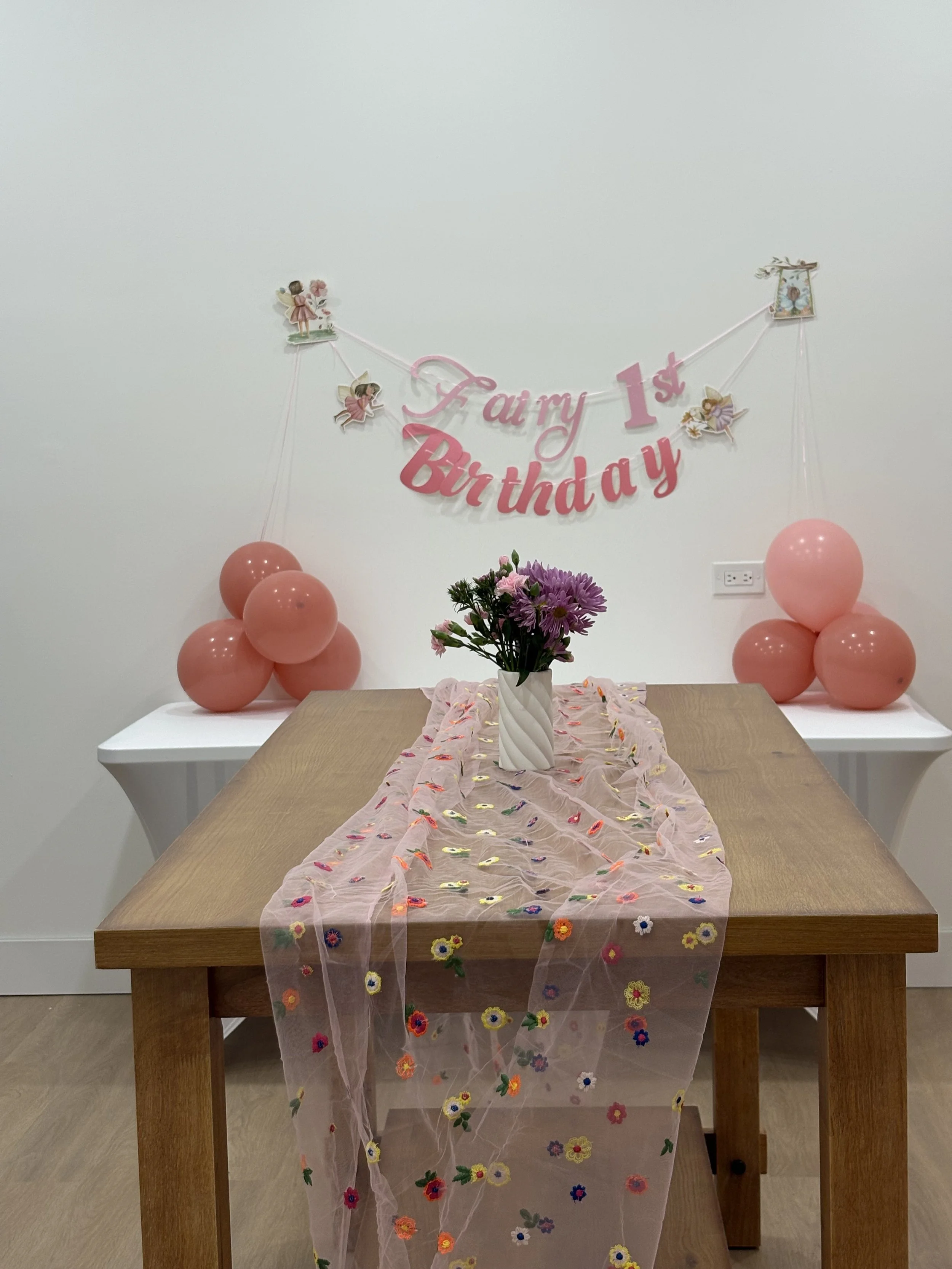 Parties table decorated for a first birthday, with pink balloons, a pink floral table runner, a vase with pink and purple flowers, and a wall banner that reads 'Fairy 1st Birthday' with fairy and bird illustrations.
