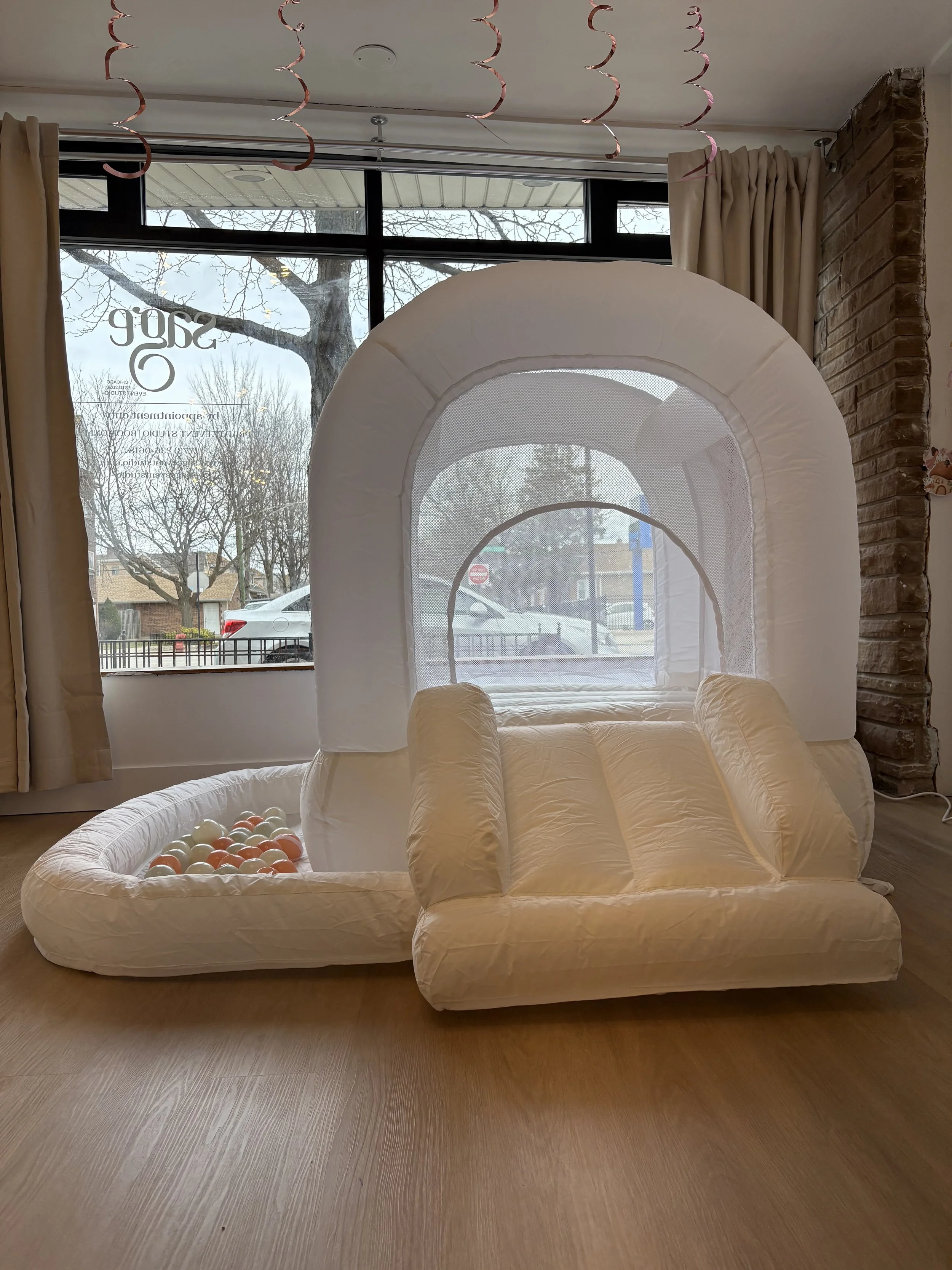 An indoor inflatable play structure with a slide, designed for children, situated near large window with curtains and tree views outside.