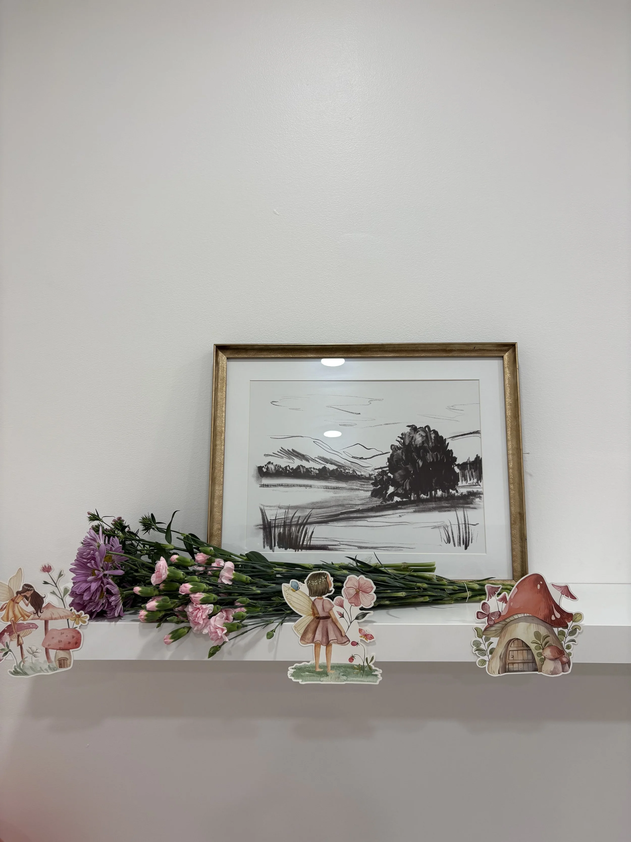 A white shelf with pink and purple flowers, fairy and mushroom cutouts, and a framed black and white landscape drawing in the background.