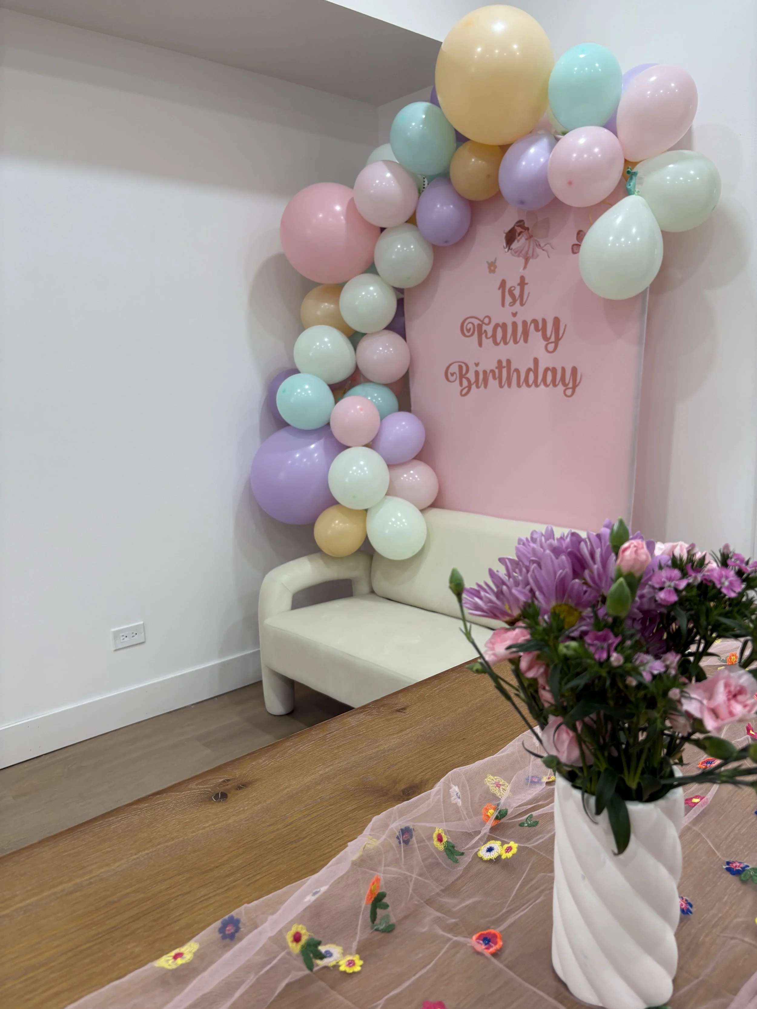 Decorative setup for a child's first fairy birthday, featuring a pink backdrop with balloons in pastel colors, a white couch, a wooden table with a vase of pink and purple flowers, and a pink tulle fabric with embroidered flowers.