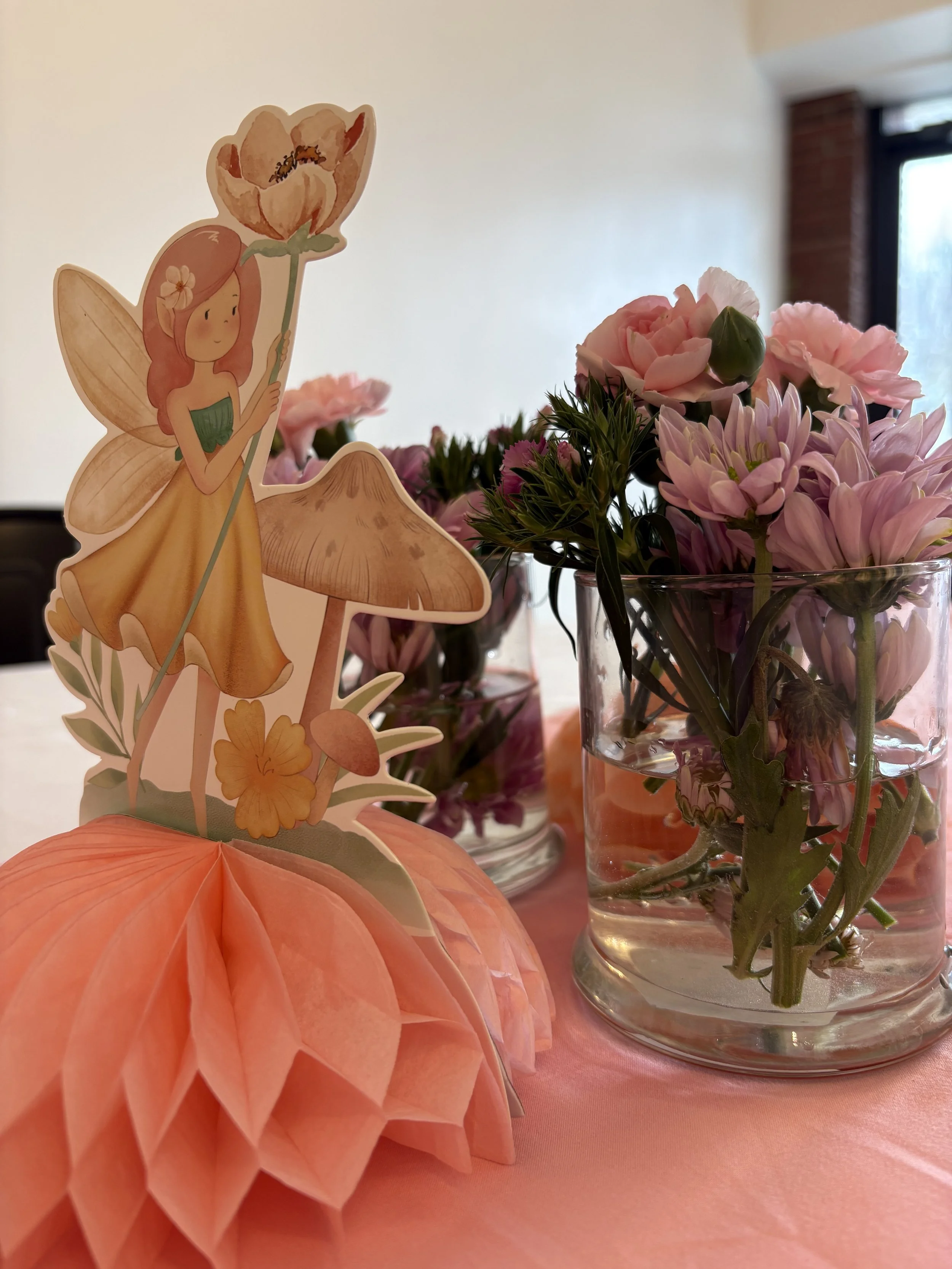 A table decorated with pink flowers in glass jars and a paper pumpkin with a fairy-themed paper figure holding a flower, set against a background of a white wall and a window.