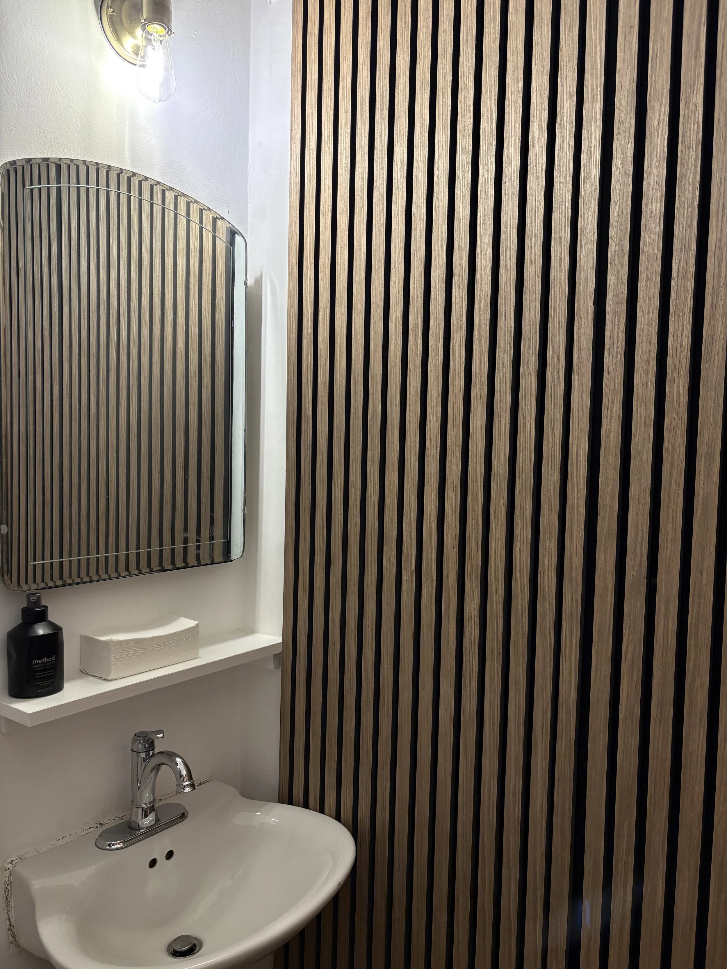 Small bathroom with a wall-mounted mirror, a small white sink with a chrome faucet, a shelf above holding a black soap dispenser and a stack of tissue paper, and a wall with vertical wooden slats.