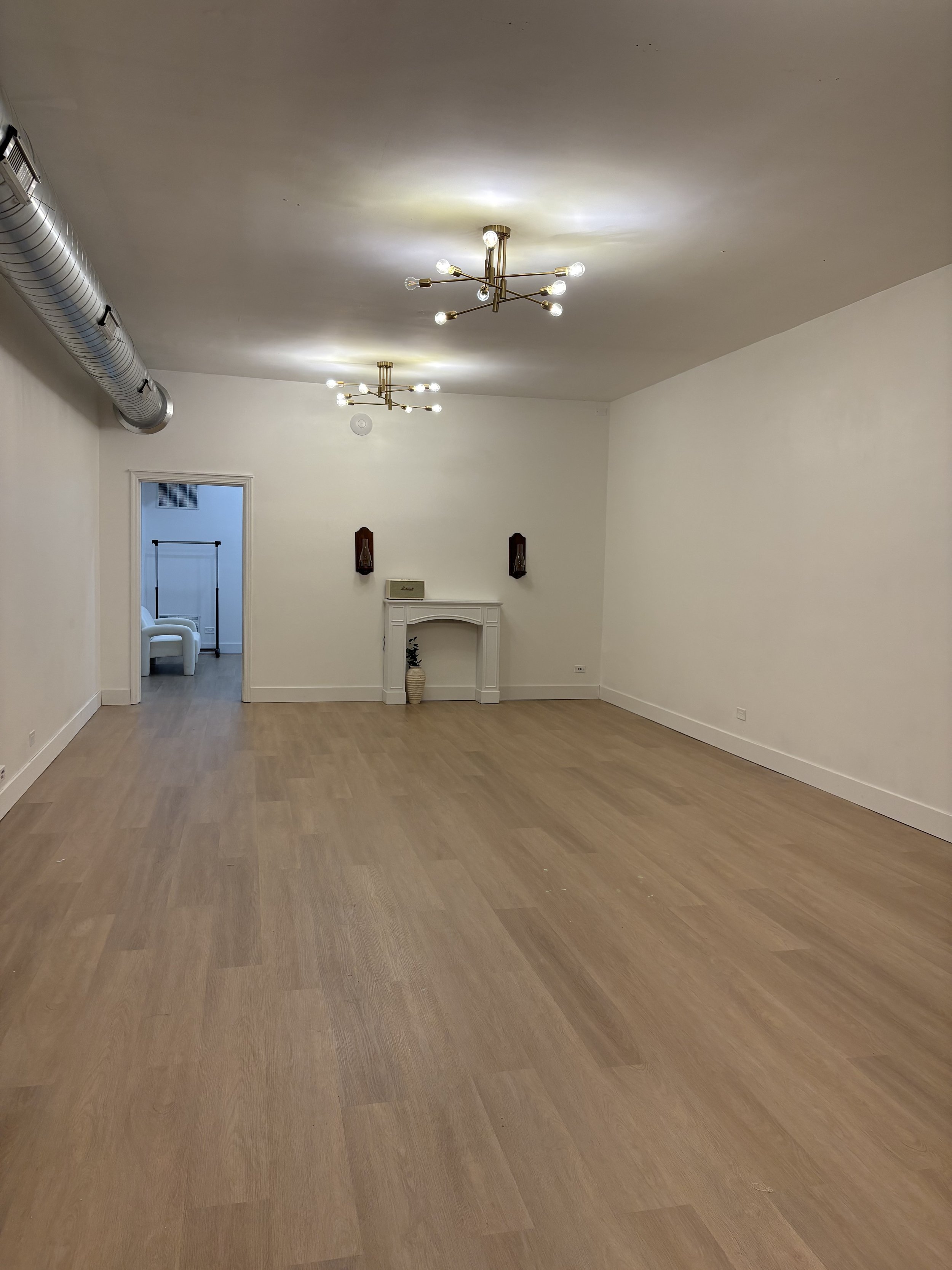 Empty living room with wooden floor, two modern chandeliers, a small fireplace with decor items, and a doorway leading to another room with a white chair.