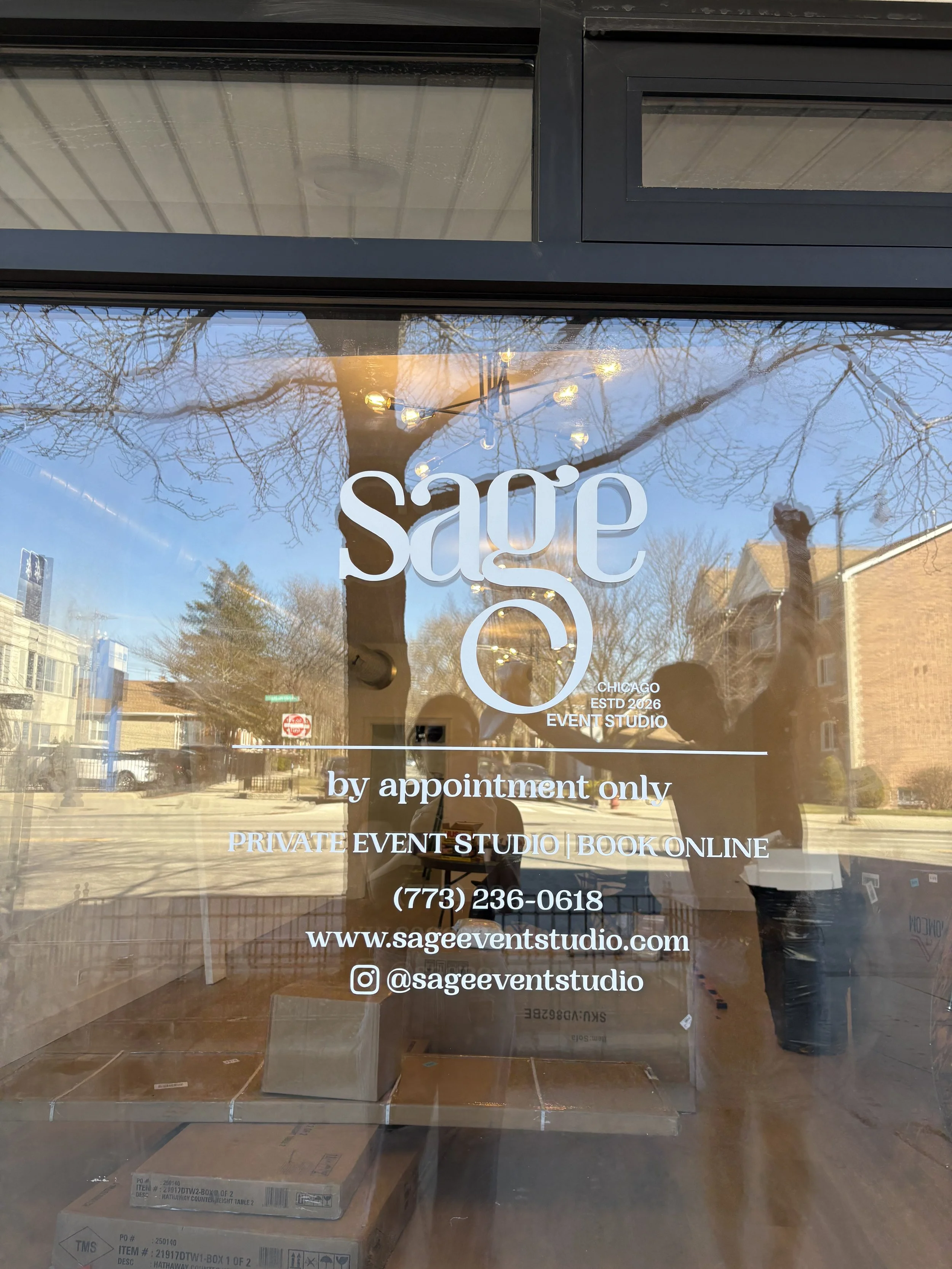 Sign on a glass door for Sage Event Studio. It states that the studio in Chicago is by appointment only, offers private event studio bookings online, and provides contact information including a phone number, website, and Instagram handle. The reflection of trees, nearby buildings, and a person taking the photo can be seen on the glass.
