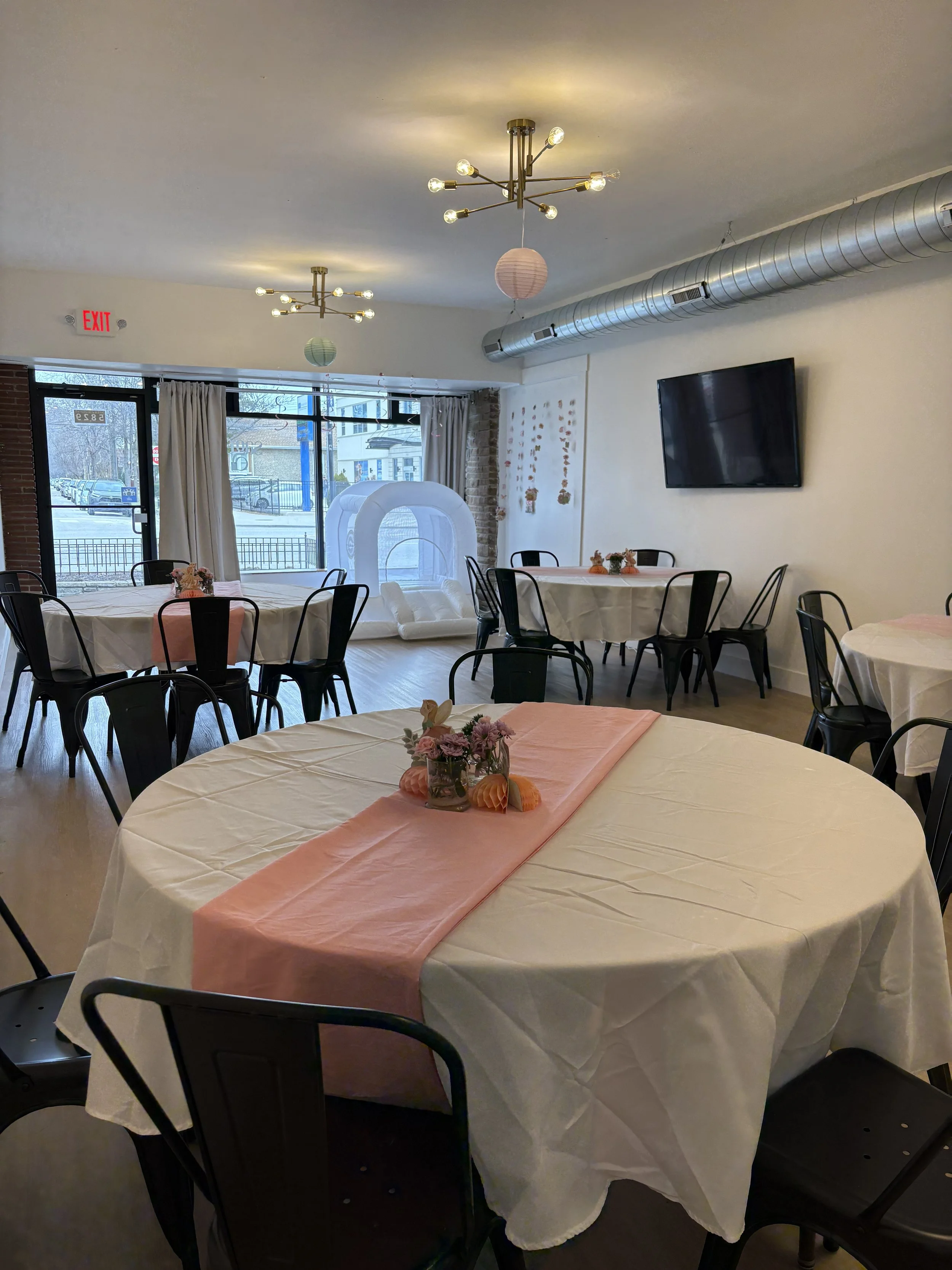 Event space decorated with round tables covered with white tablecloths and pink table runners, floral centerpieces, and black chairs. Large windows with white curtains, a wall-mounted TV, modern ceiling lights, and paper lanterns. An inflatable bounce house inside the room.