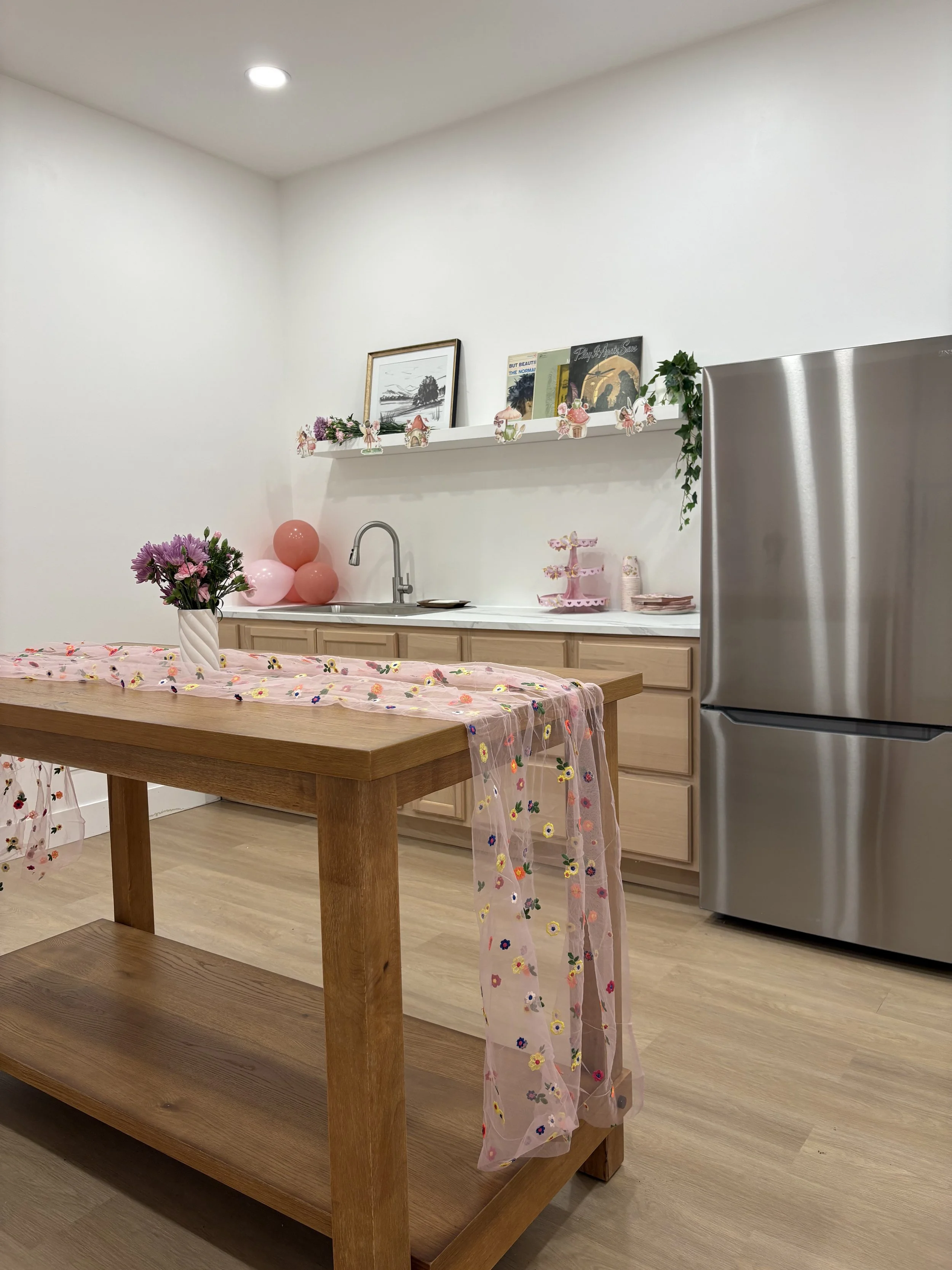 Decorated kitchen with pink and purple flowers, balloons, and a sheer pink table runner with embroidered flowers, white walls, and minimal decor.