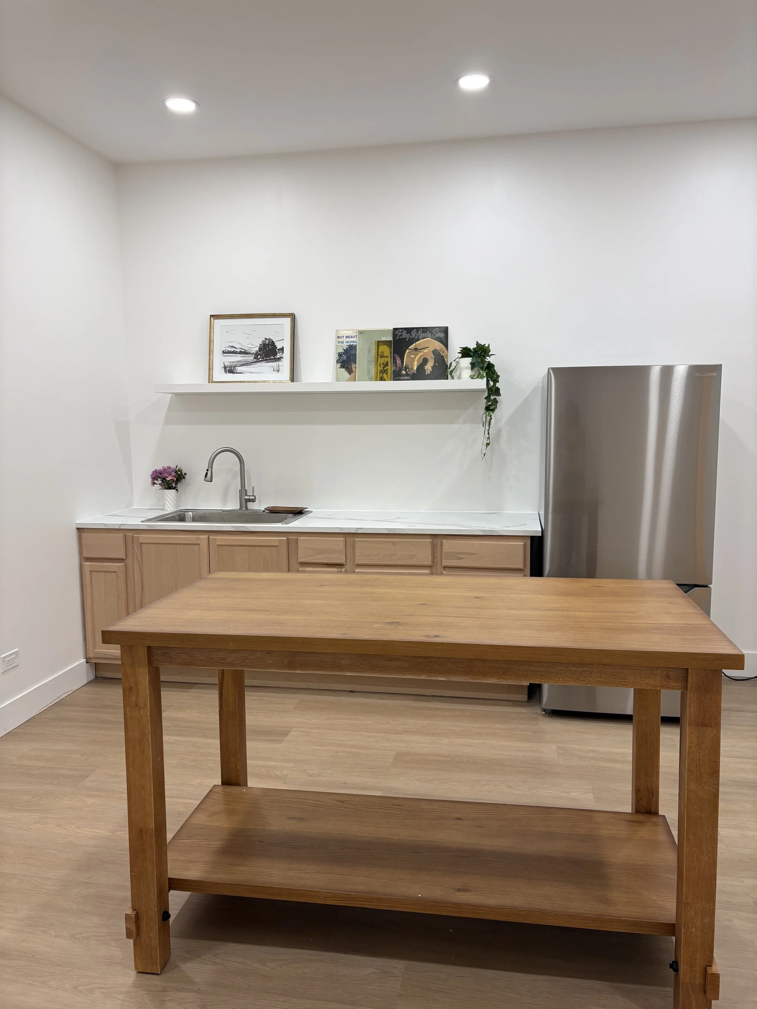 A minimalist kitchen with a wooden island, light wood cabinets, a stainless steel refrigerator, and a white shelf with framed pictures, books, a green plant, and a small bouquet of purple flowers.