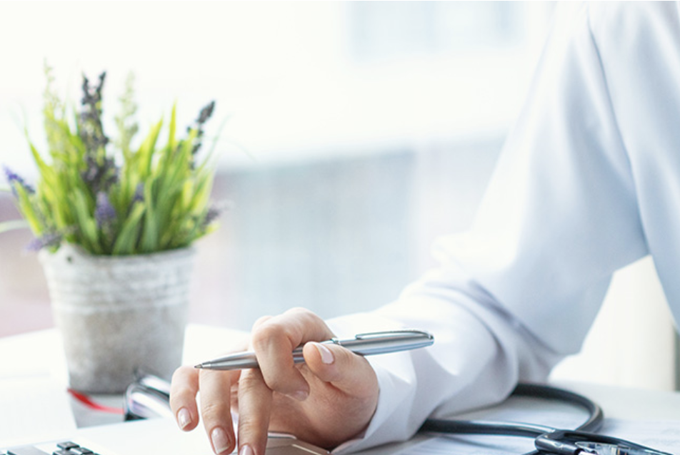 A person in a white coat holding a pen, seated at a desk with a stethoscope, with a potted plant in the background.