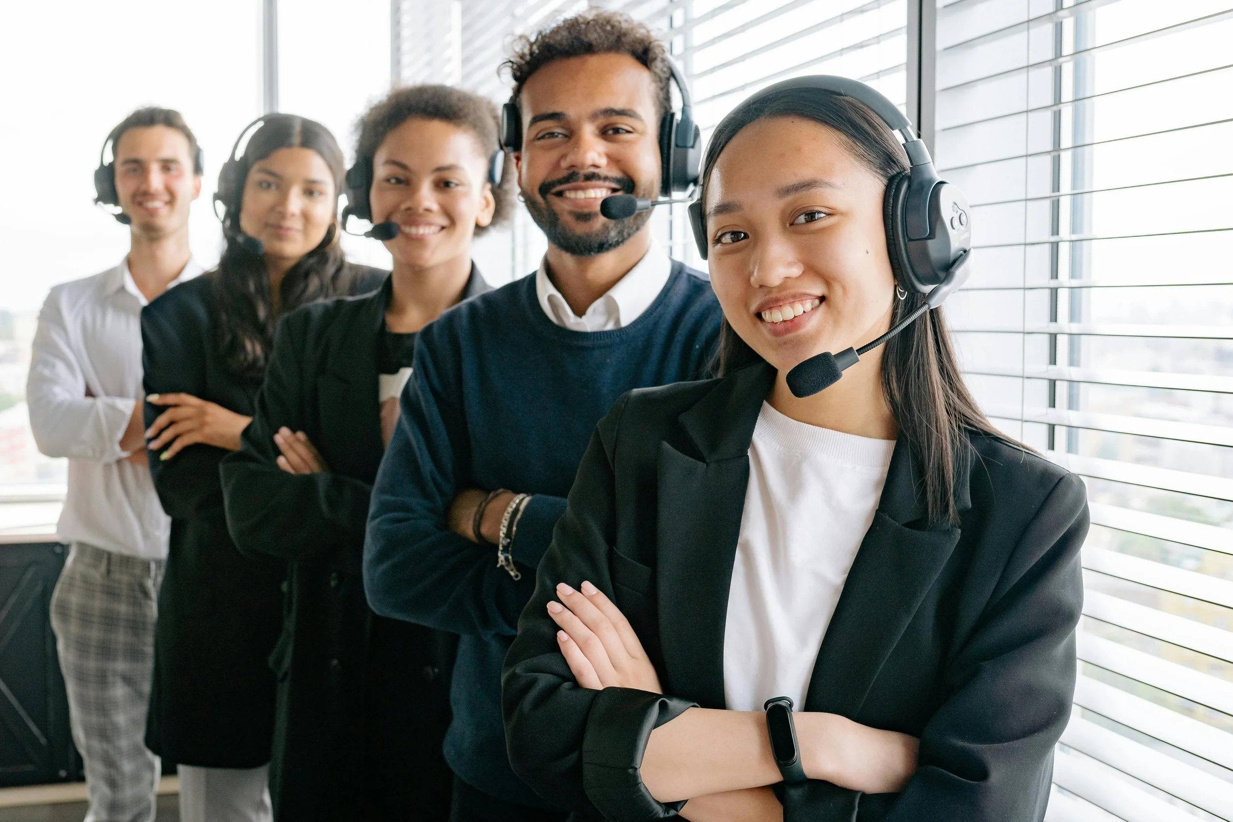 Team of five customer service representatives standing in a line, smiling, wearing headsets, with a large window in the background.