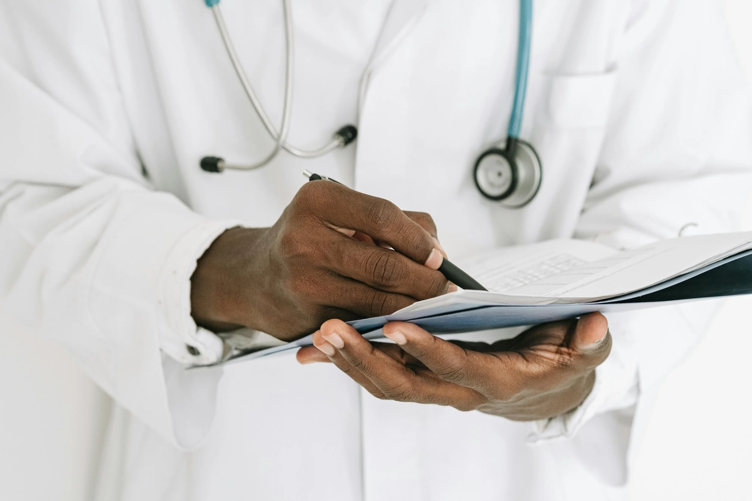 A healthcare professional, wearing a white lab coat and stethoscope, is holding a clipboard and writing with a pen.
