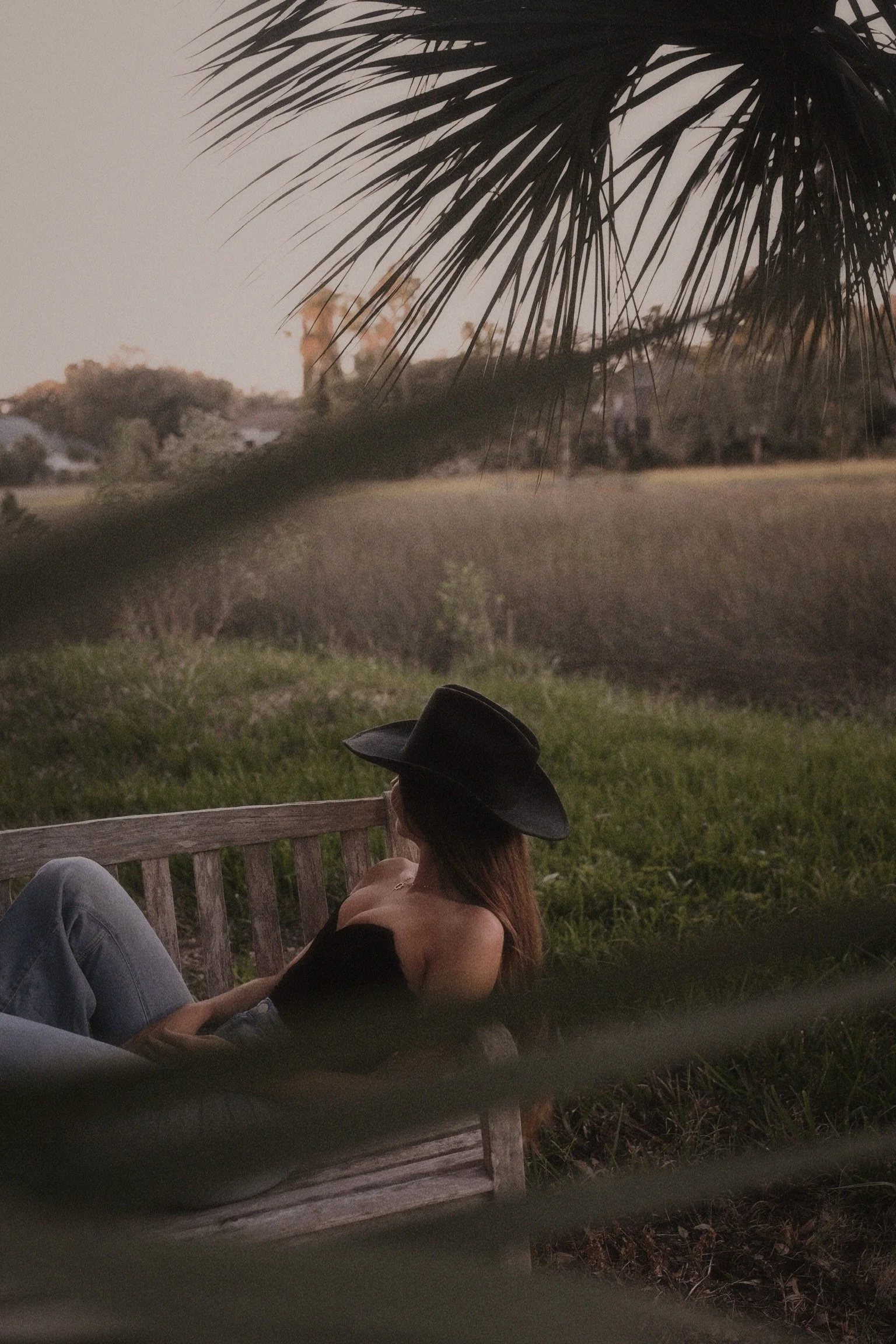 A woman with red hair, wearing a black hat and black top, is sitting on a wooden bench in a grassy area, looking at a rural landscape during sunset, with trees and fields in the background, partially obscured by a palm leaf in the foreground.