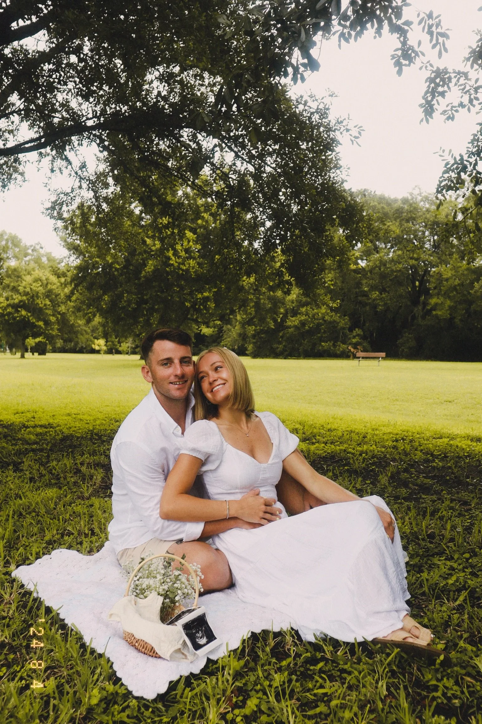 A happy couple sits on a picnic blanket in a park, surrounded by green grass and trees, with a basket and ultrasound photo nearby.