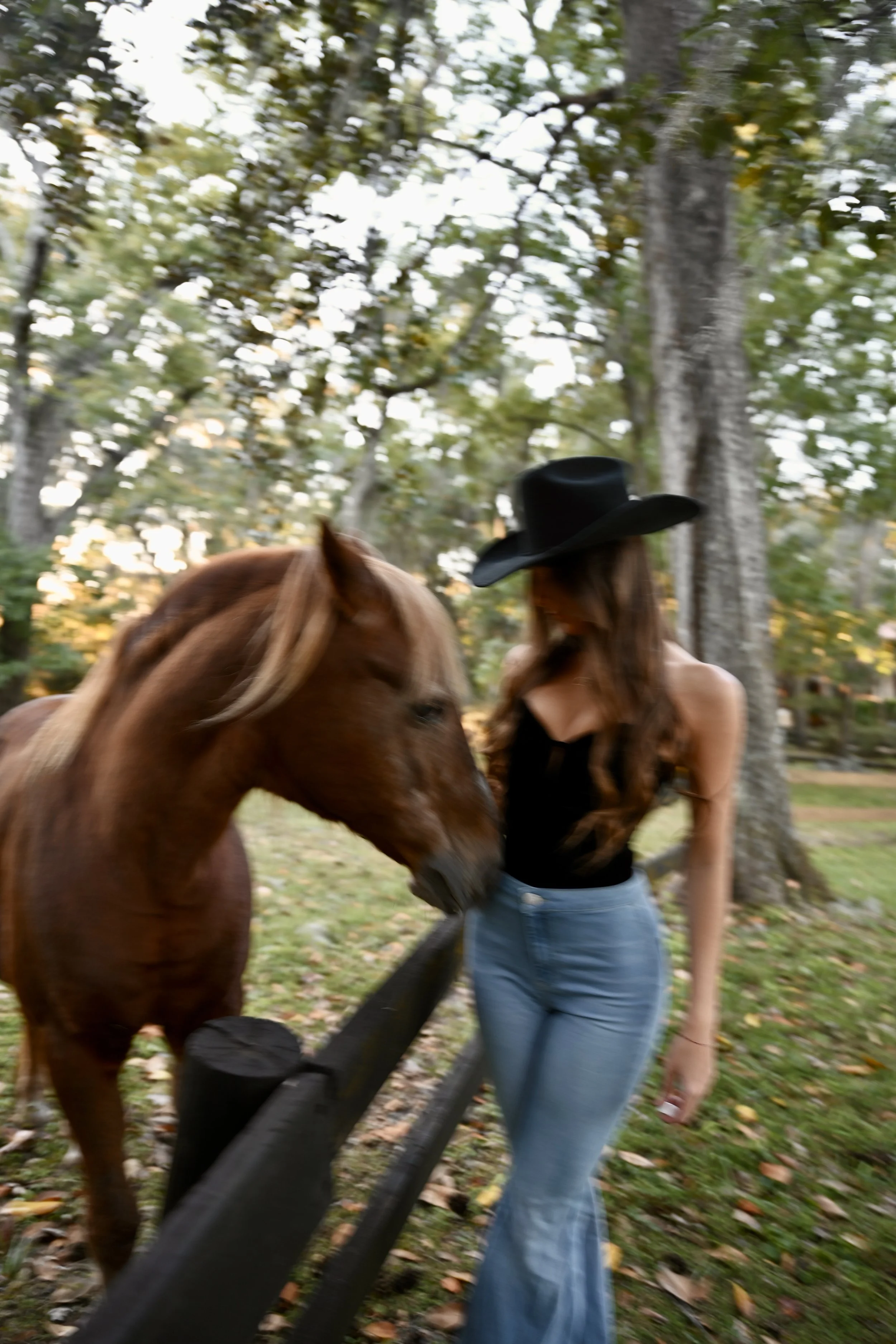 A woman with long hair in a black top, standing next to a brown horse near a wooden fence in a wooded area.