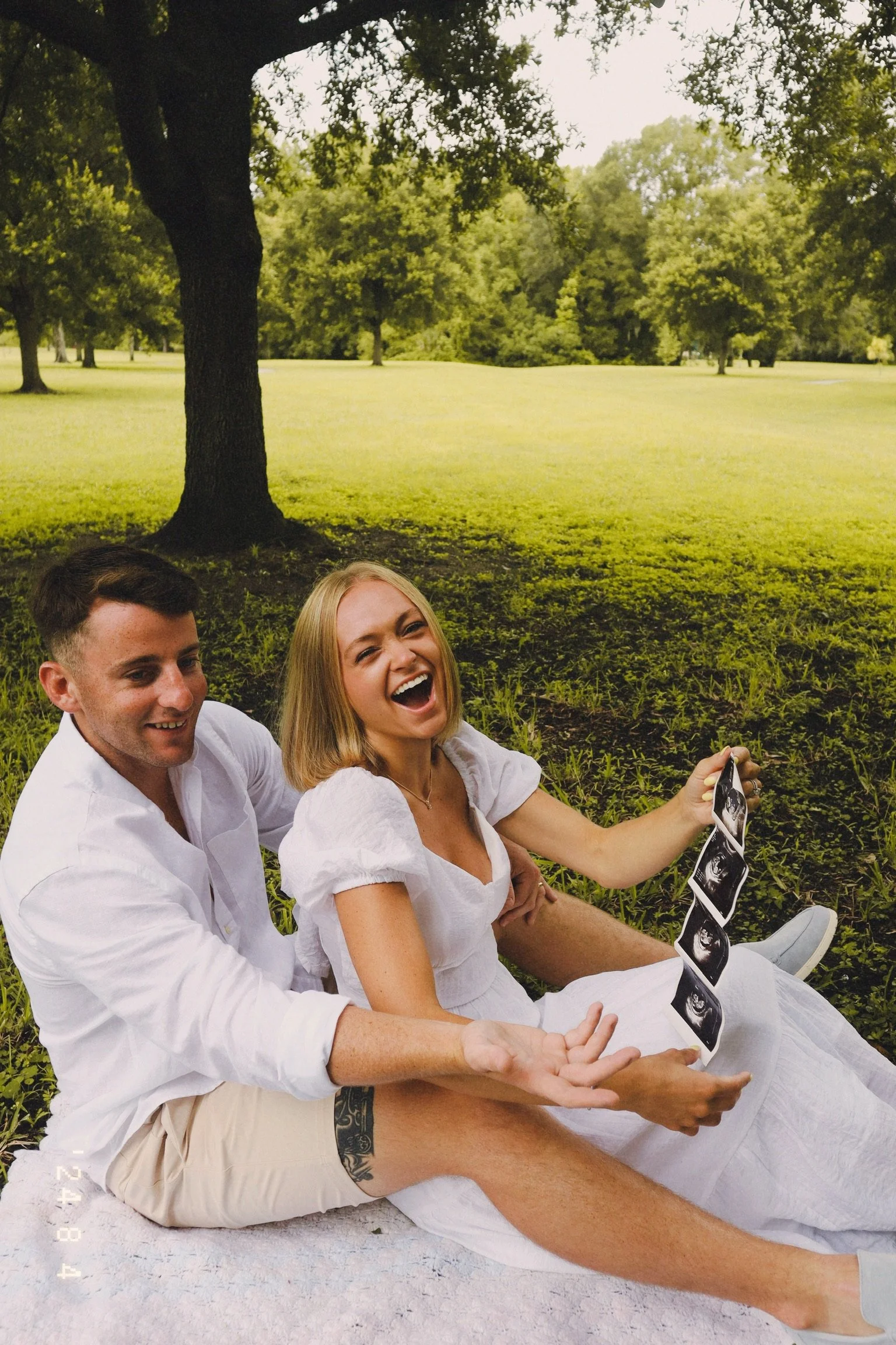 A smiling woman and a man sitting on a blanket under a large tree in a park, holding ultrasound images and sharing a joyful moment.