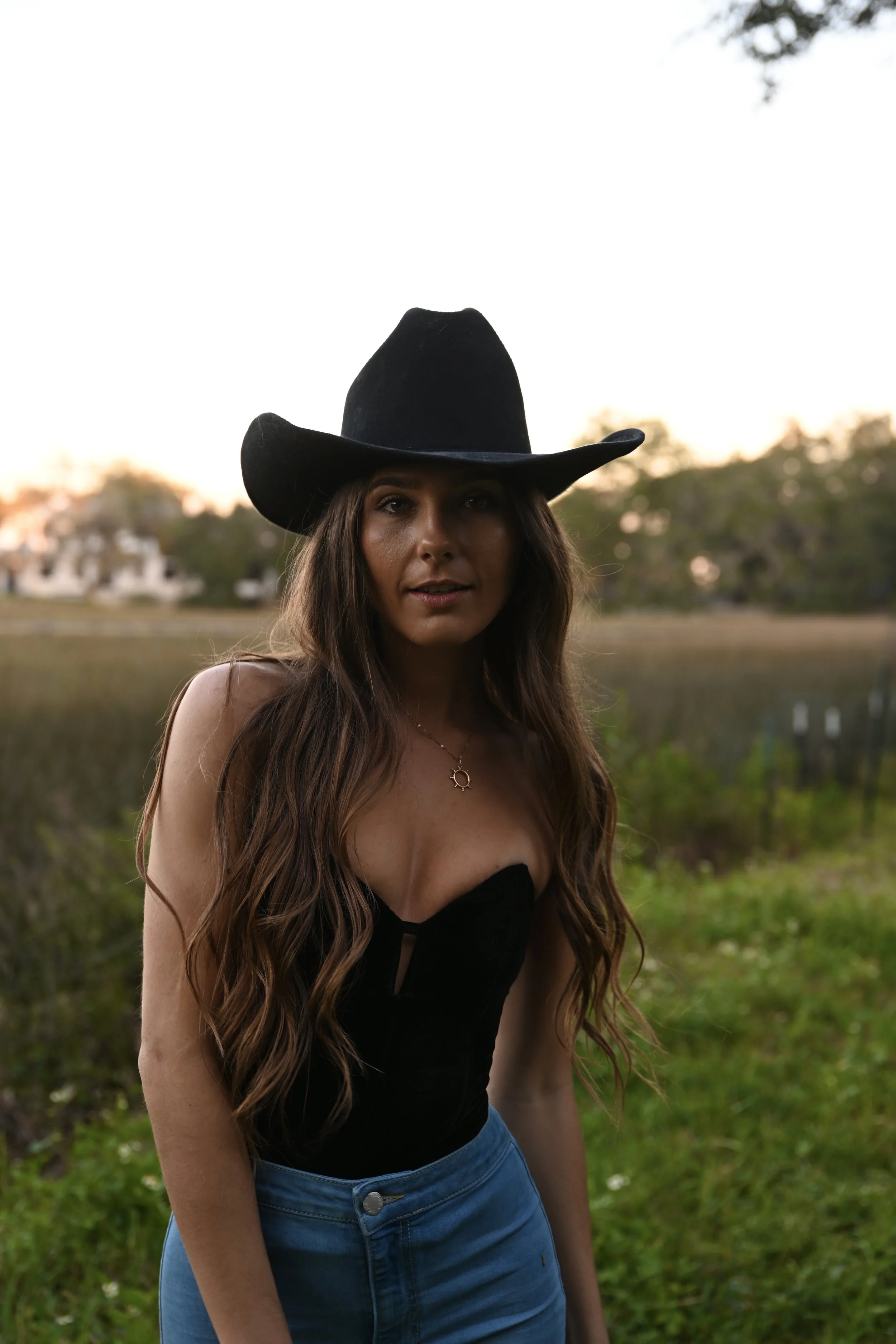 A woman wearing a black cowboy hat, black strapless top, and blue jeans outdoors during sunset.