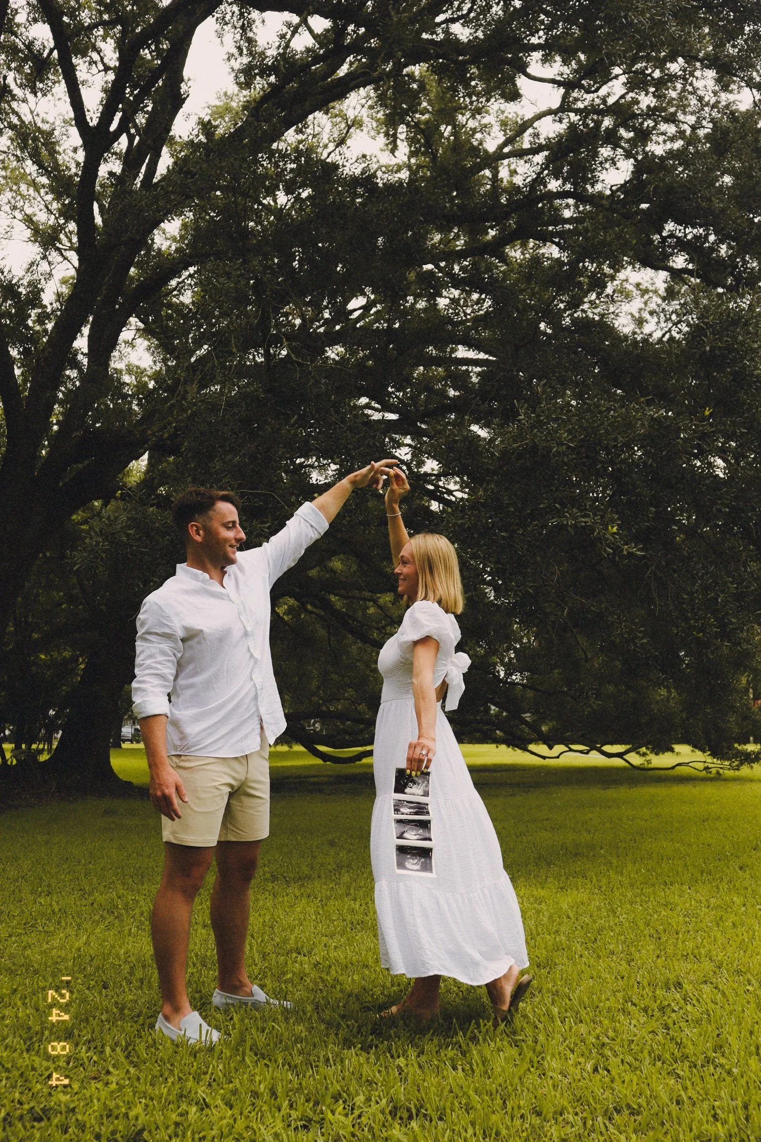 A couple stands on green grass in front of a large tree, with the man twirling the woman as she holds ultrasound images in her hand.