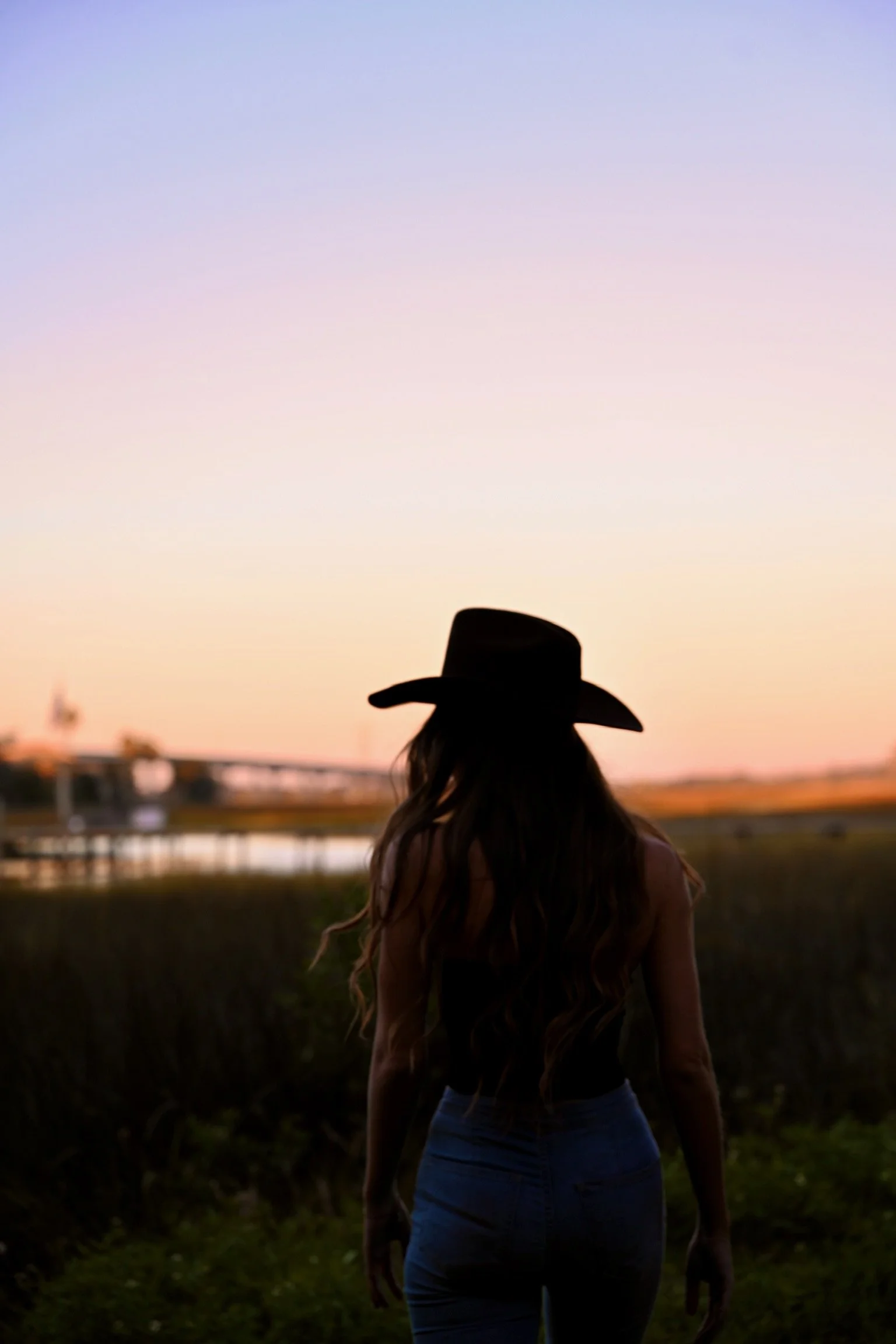 Silhouette of a woman with long hair wearing a cowboy hat, standing outdoors during sunset or sunrise.