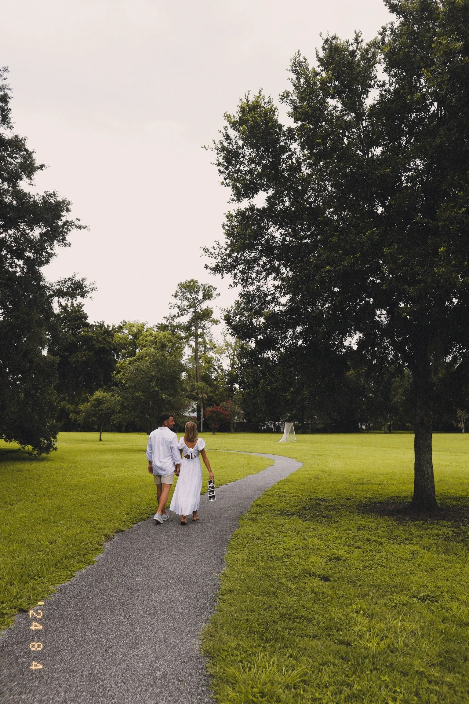 A couple walking hand in hand on a winding path through a park, surrounded by green grass and trees, with an outdoor netted sports goal in the background.