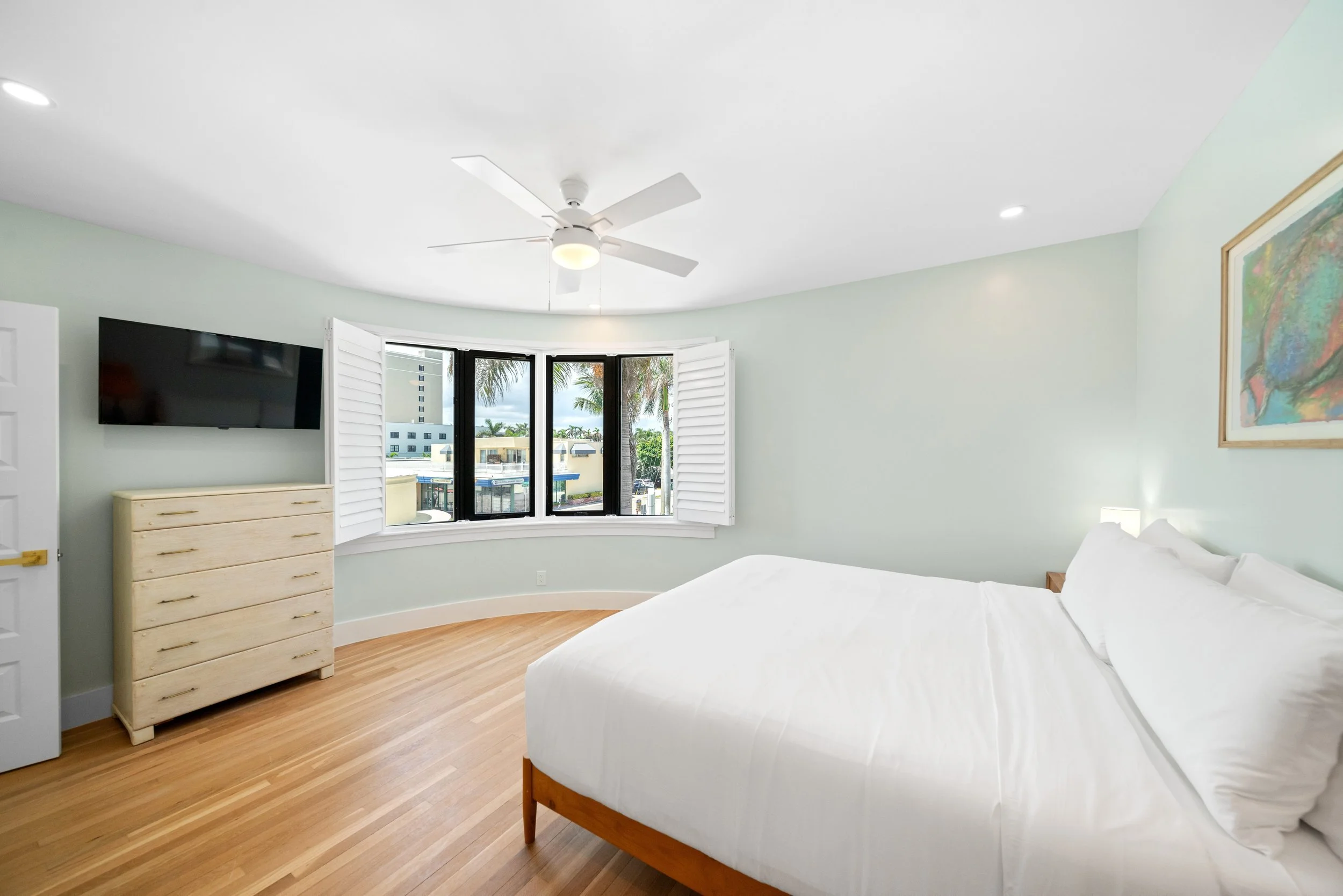 Bright bedroom with a large window, white bed, dresser, tv, ceiling fan, hardwood floors, and artwork on the wall.
