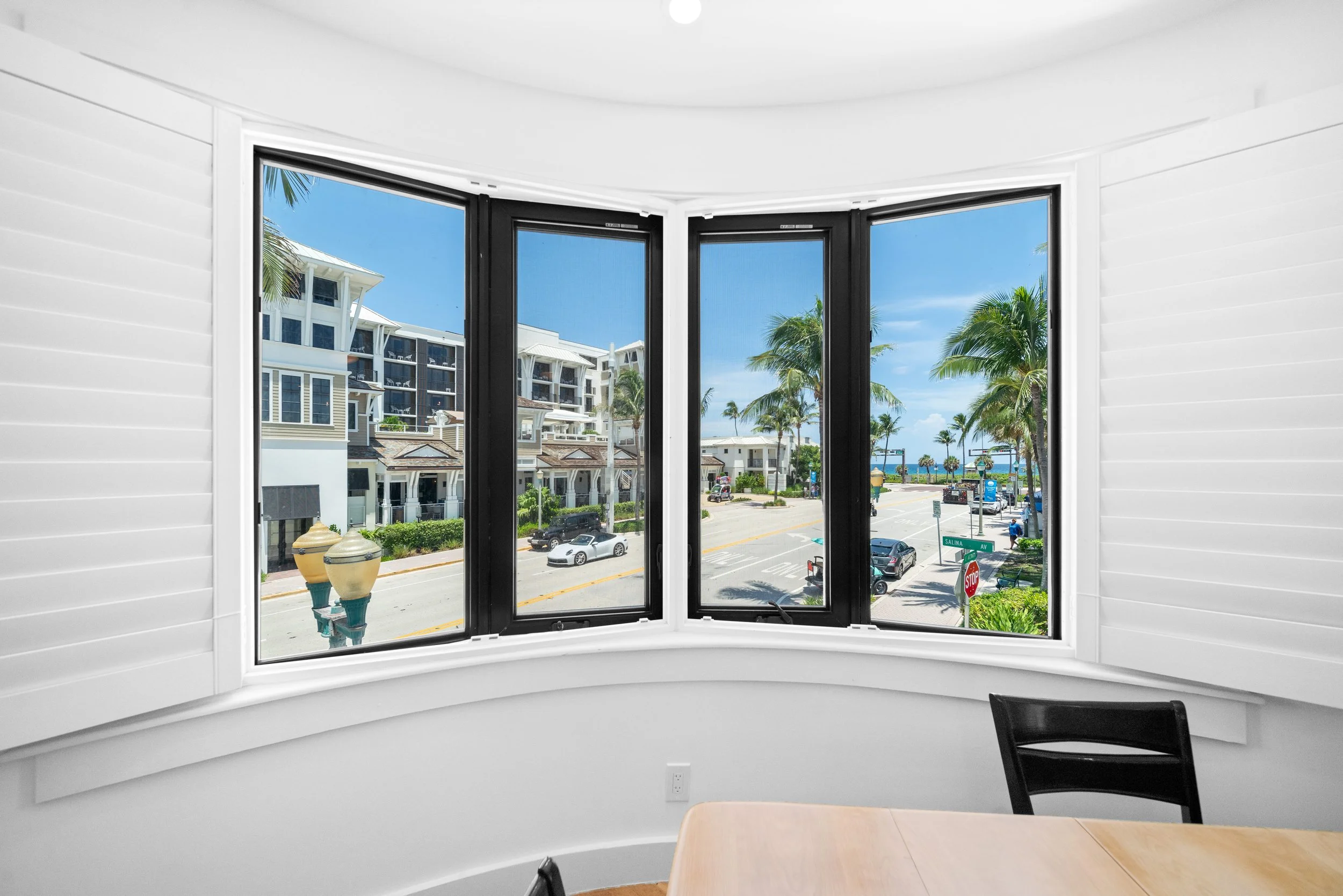 View of a street with palm trees and modern buildings through a large bay window in a white room.