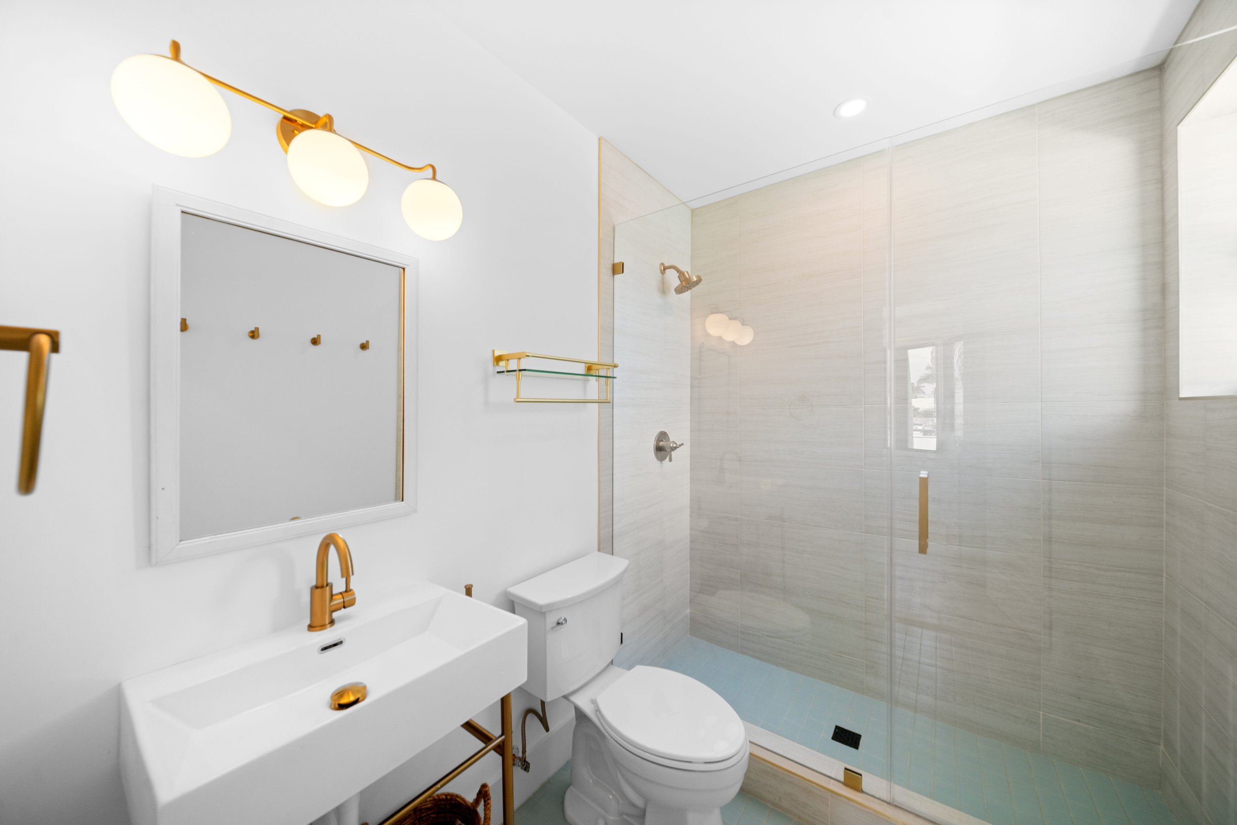 Modern bathroom with white walls, gold fixtures, a white sink with gold accents, a mirror, a toilet, and a glass-enclosed shower with beige tiles and a gold showerhead.