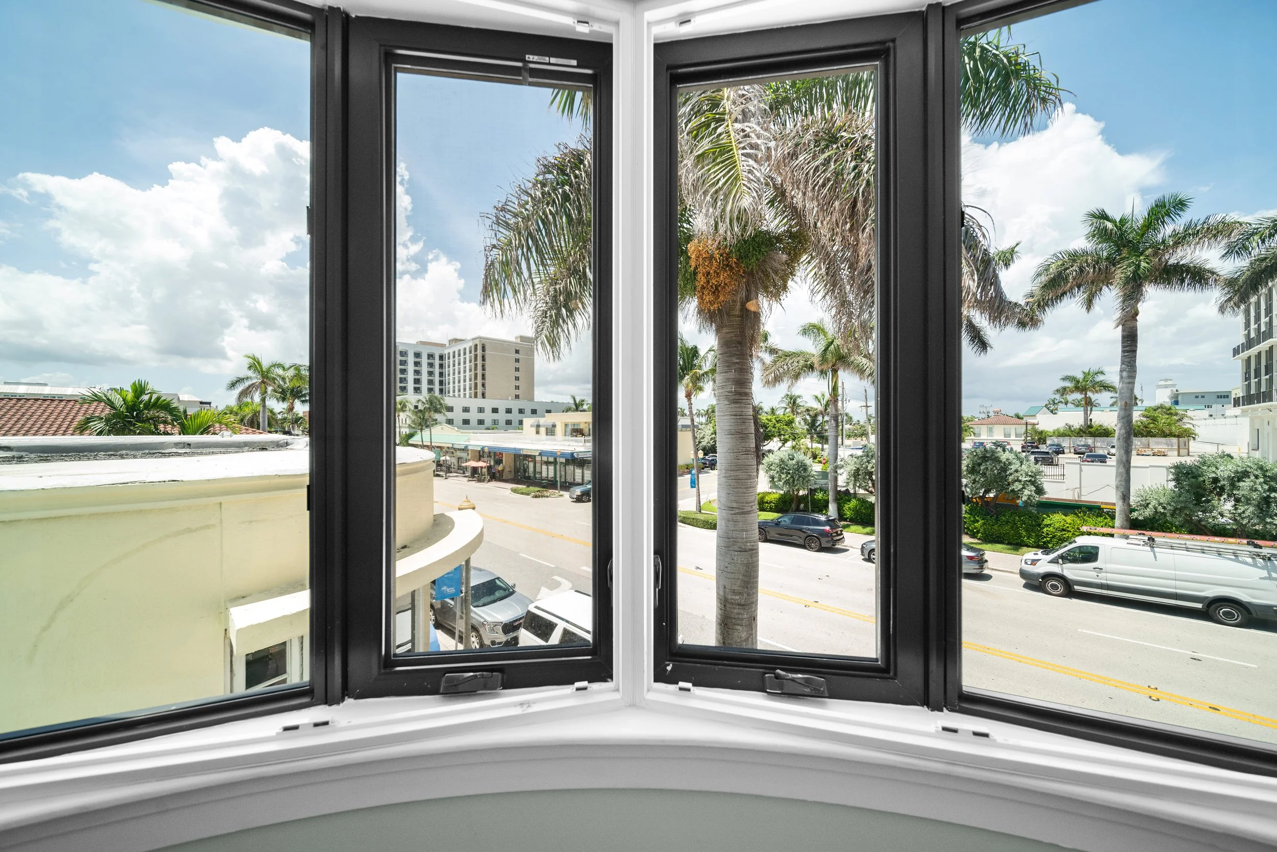 View of a street with palm trees and cars through a bay window with black frames in a bright, sunny day.
