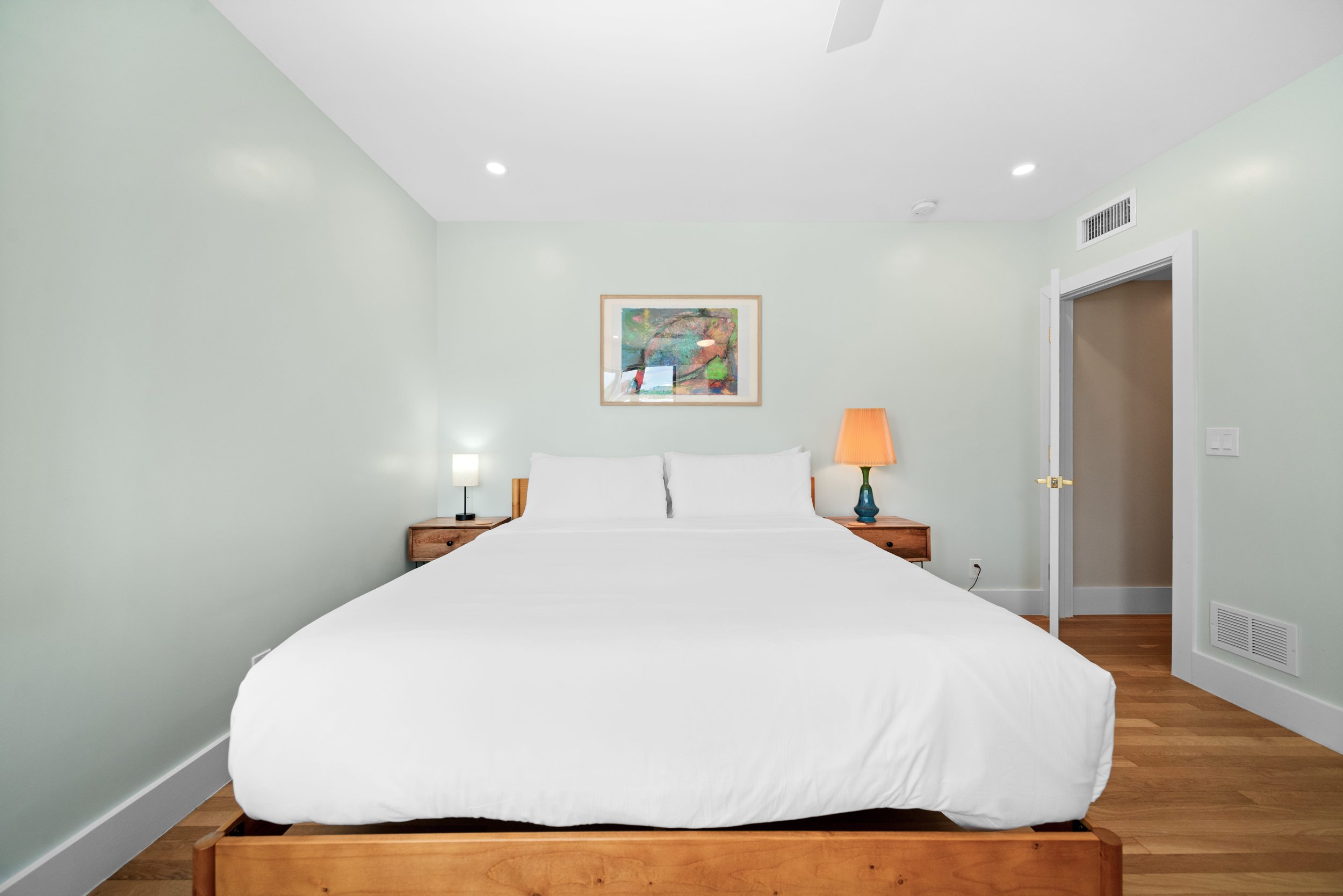 A neatly made bed with white sheets and pillows, two wooden bedside tables with lamps, a framed artwork above the bed, and an open door in a light-colored bedroom with hardwood floors.
