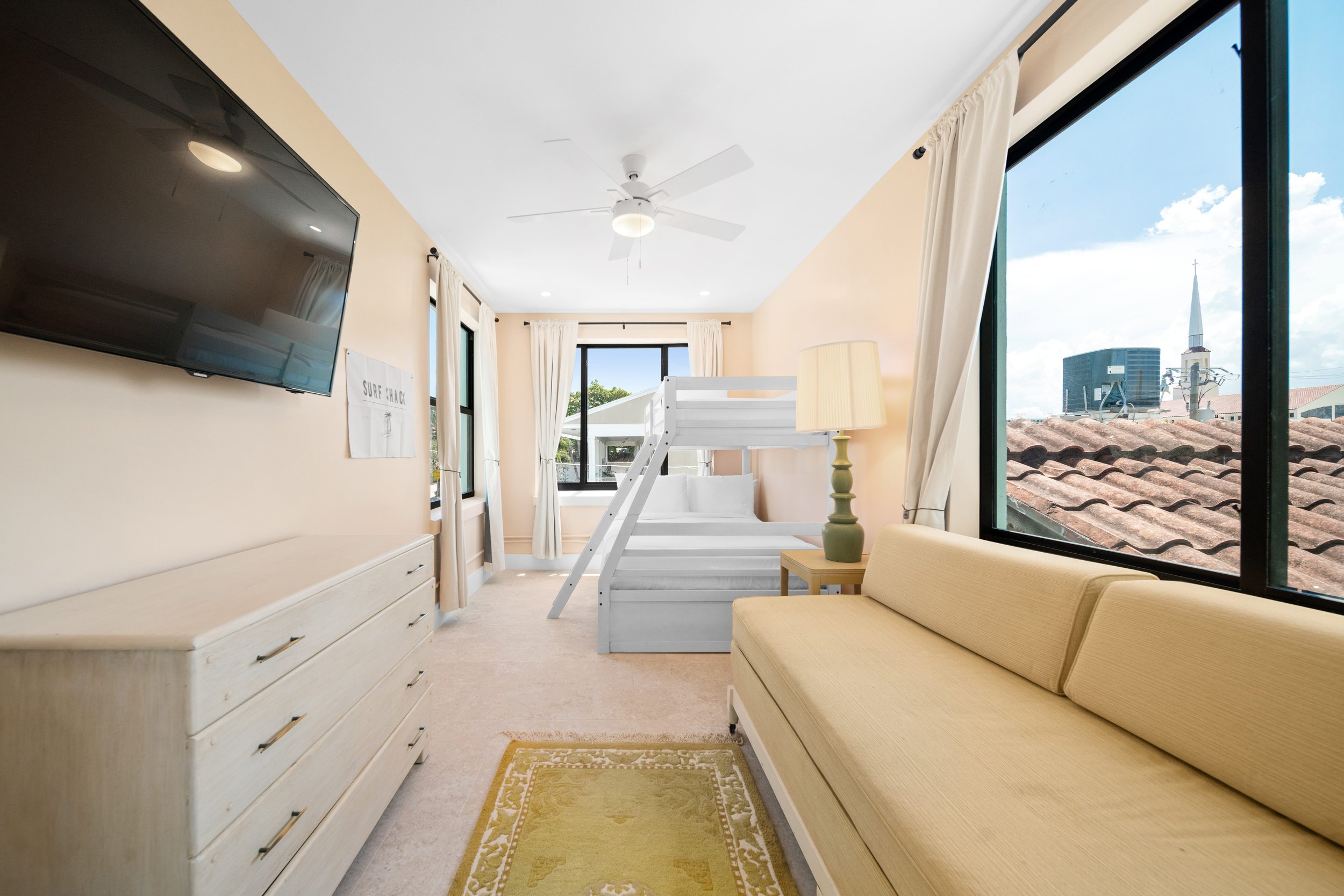 Bright hotel room with large windows, beige walls, a white dresser, a yellow sofa, a lamp on a side table, and a bunk bed with white bedding. Outside the window, rooftops and city buildings are visible under a blue sky.