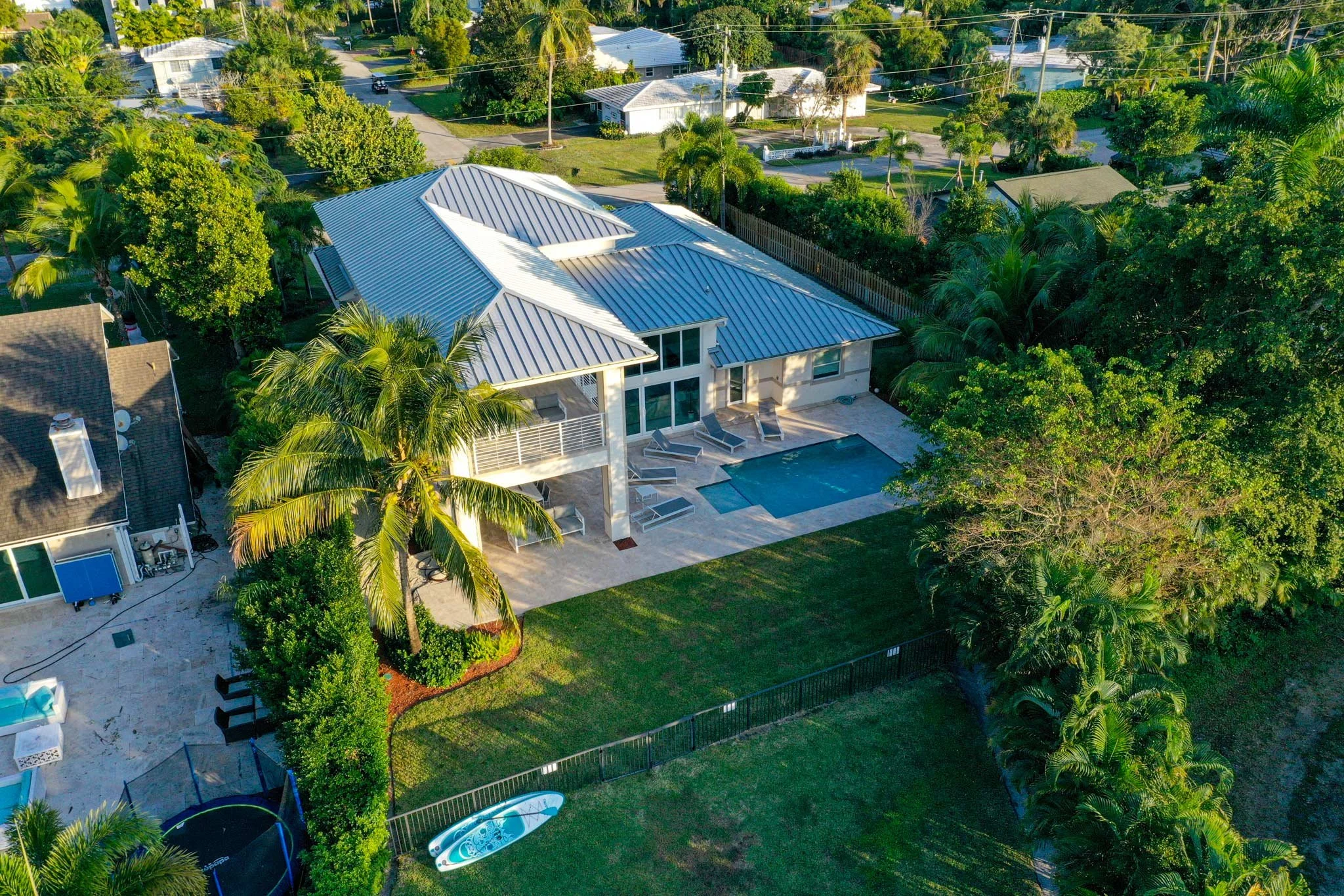 Aerial view of a backyard with a swimming pool, patio area, lawn, and palm trees surrounded by houses and lush greenery.